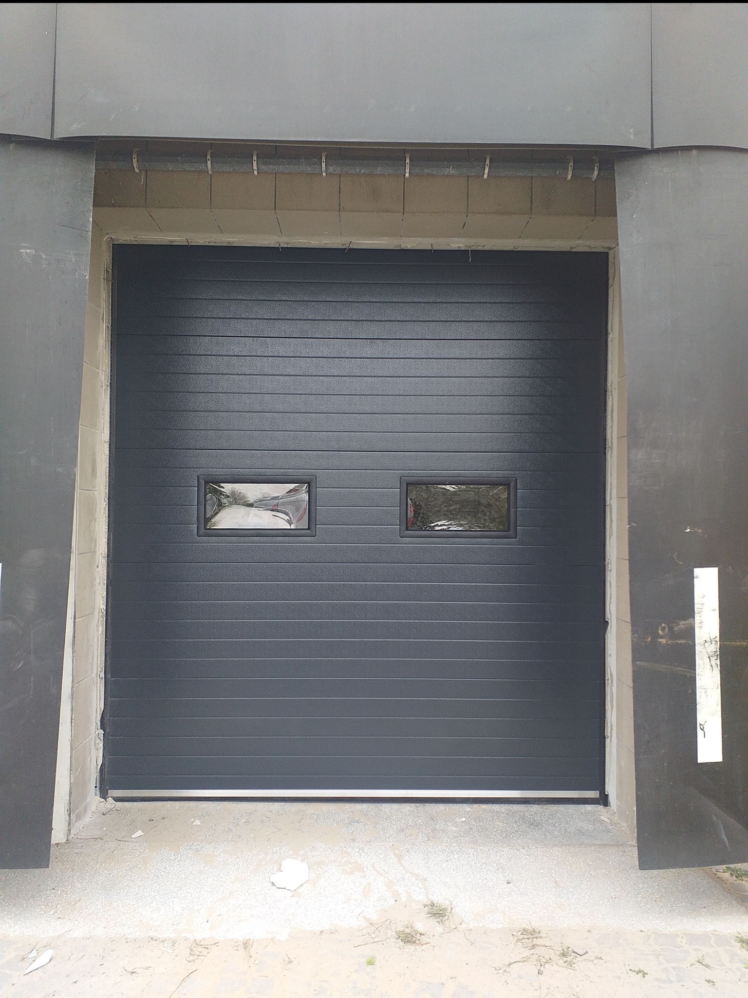Zamknięta, szara, segmentowa brama garażowa z dwoma małymi, prostokątnymi oknami, widoczna od frontu, z surowym wykończeniem murarskim po bokach.