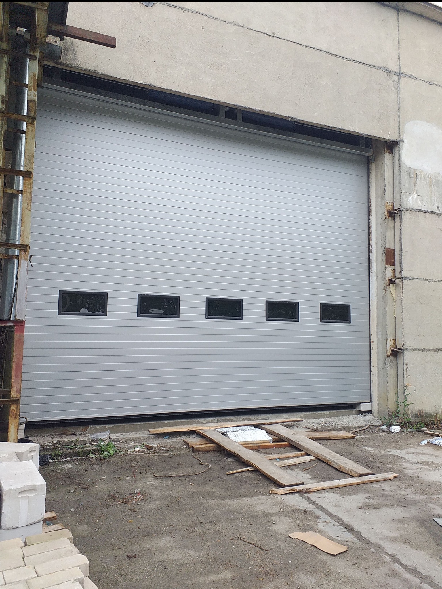 Szara segmentowa brama przemysłowa z czterema małymi okienkami, zamontowana w surowym, betonowym budynku przemysłowym, z drewnianymi elementami na ziemi przed nią.