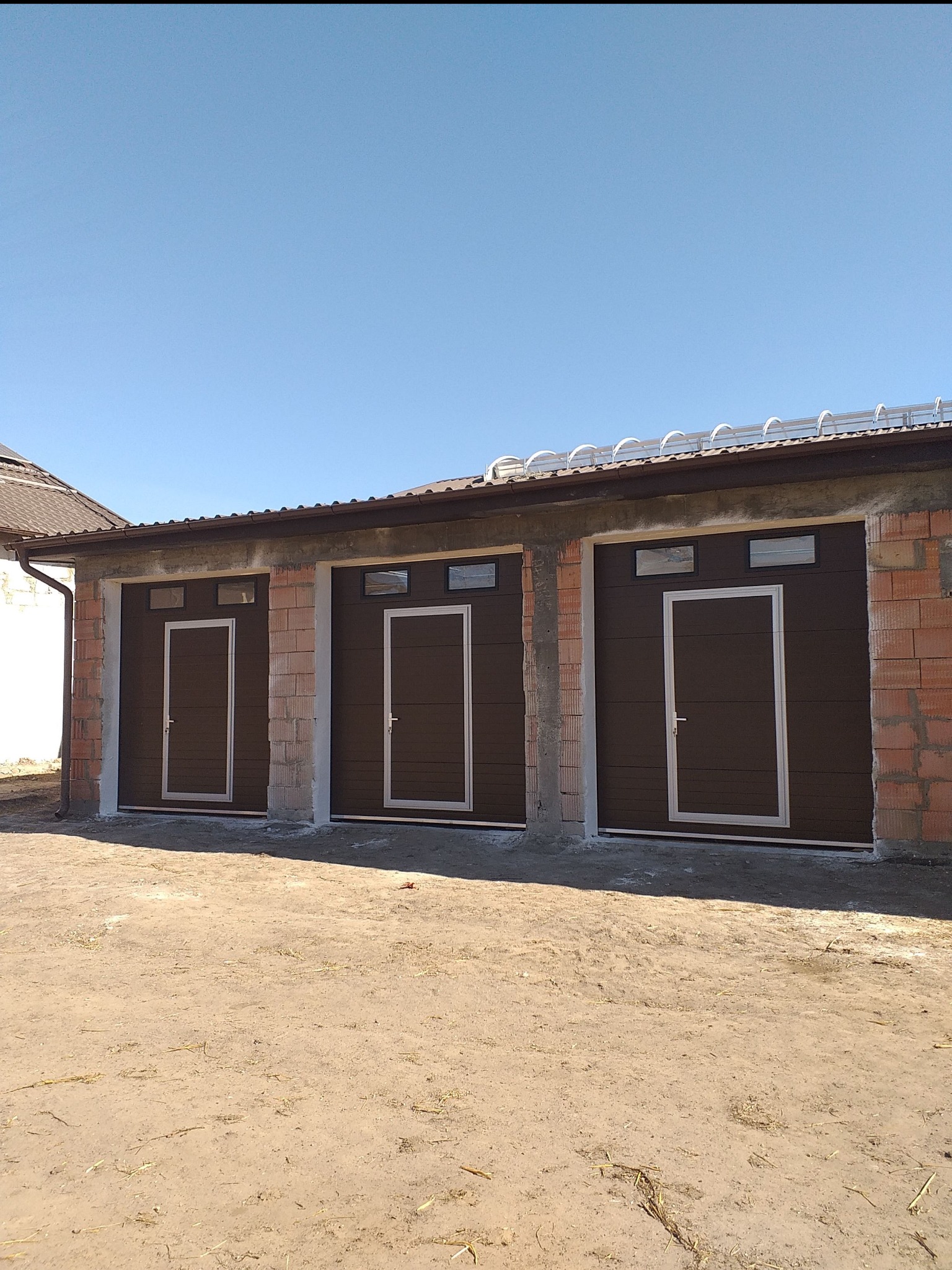 Trzy brązowe segmentowe drzwi garażowe z oknami i jasnymi ramami, zamontowane w surowych murach budynku garażowego w słoneczny dzień.