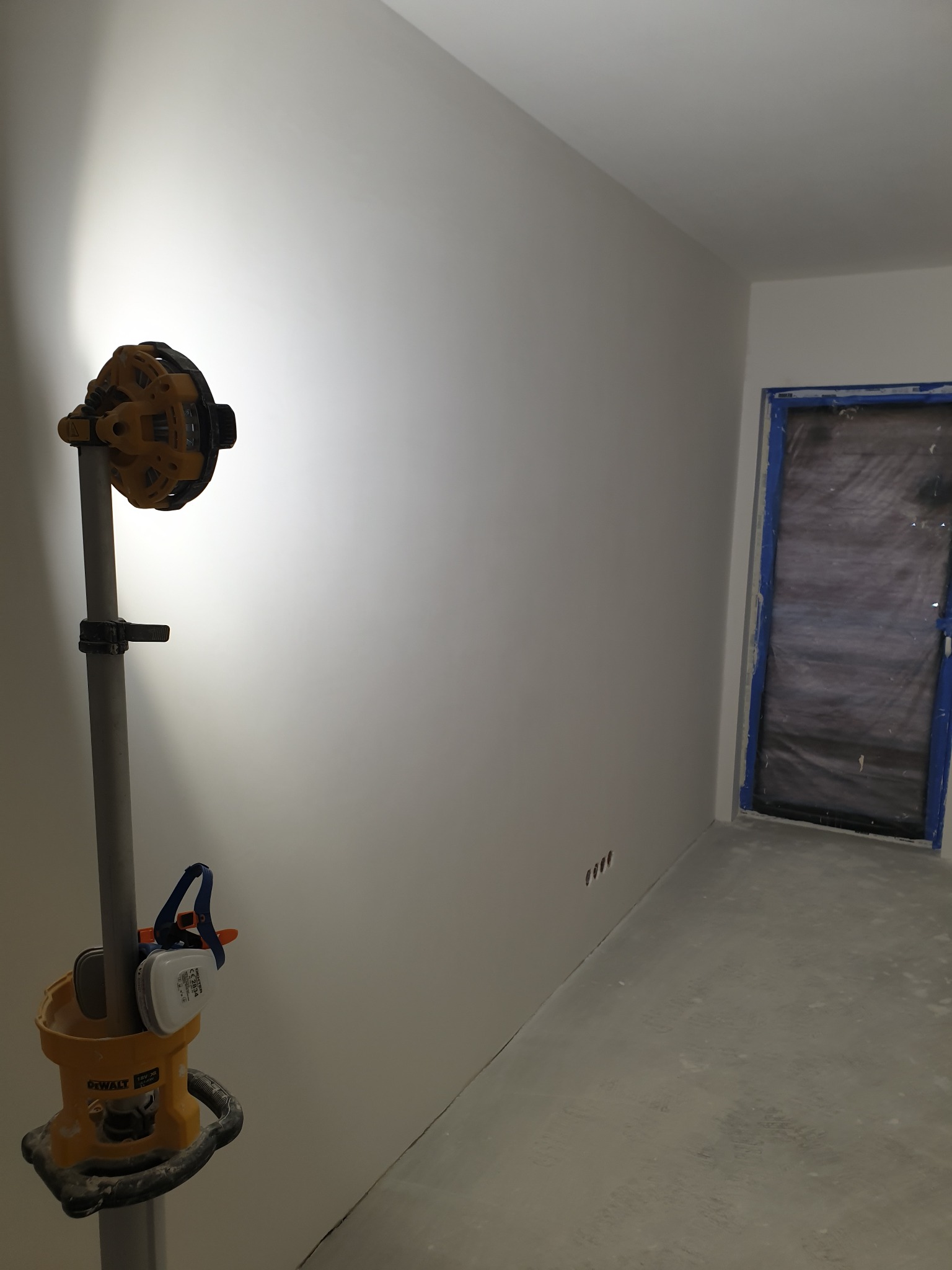 Świeżo wyszpachlowana, gładka ściana w pokoju, oświetlona lampą roboczą. Widoczne zabezpieczone folią i taśmą drzwi oraz szlifierka do gładzi na statywie.
