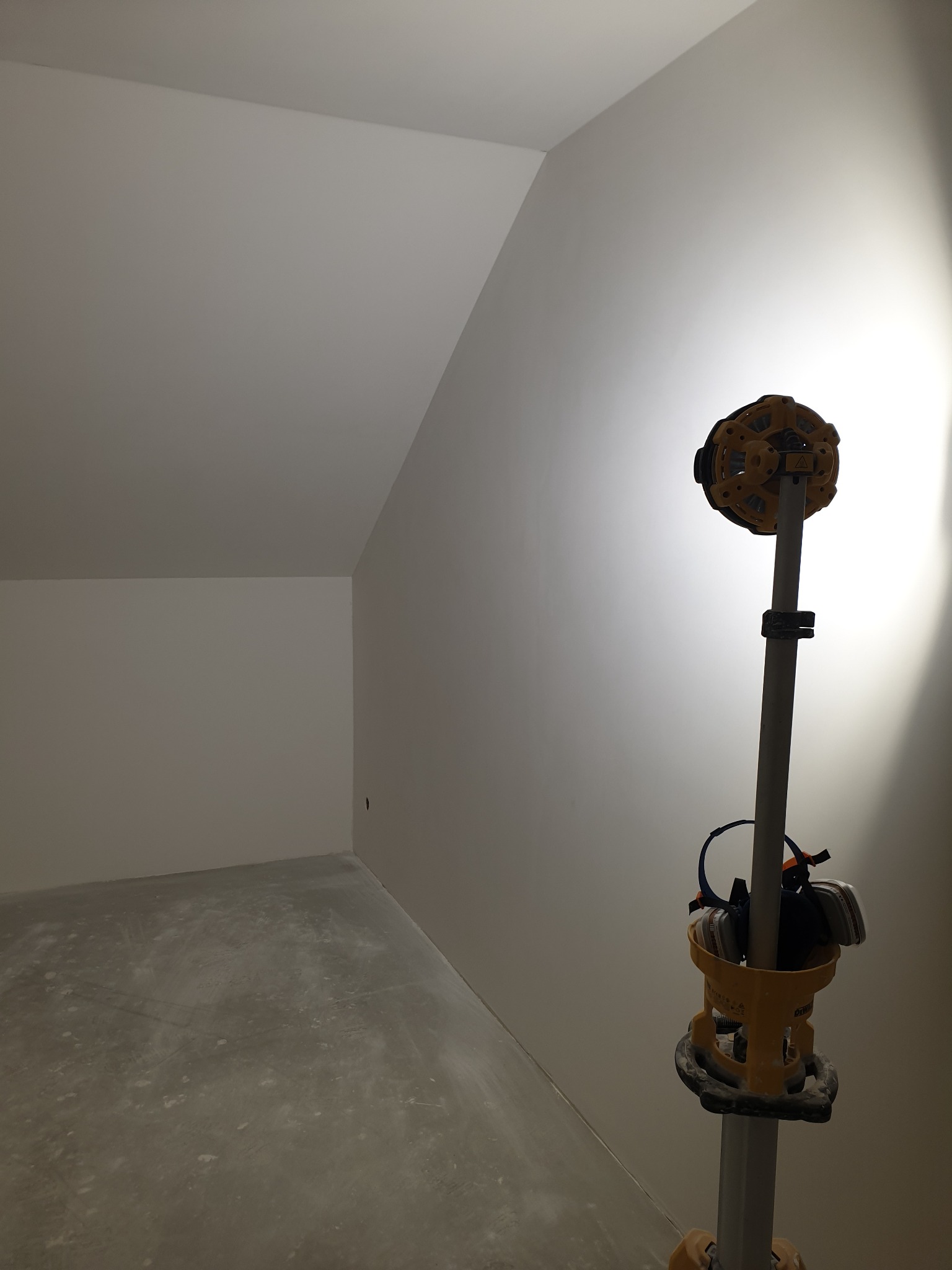 Poddasze po szpachlowaniu, widoczna gładka ściana i narzędzie do szlifowania na statywie. Surowe wykończenie podłogi. Oświetlenie punktowe.