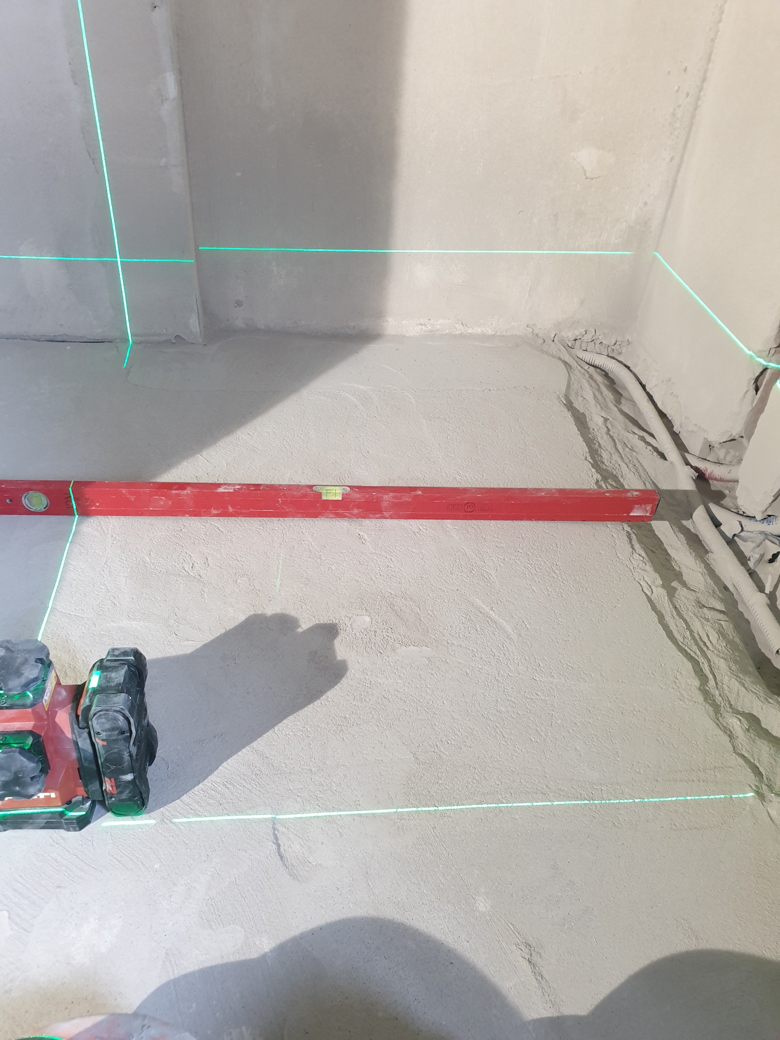 Ściana w trakcie prac wykończeniowych z poziomnicą laserową, czerwoną poziomicą i widocznymi przewodami. Zielone linie lasera wyznaczają poziom.