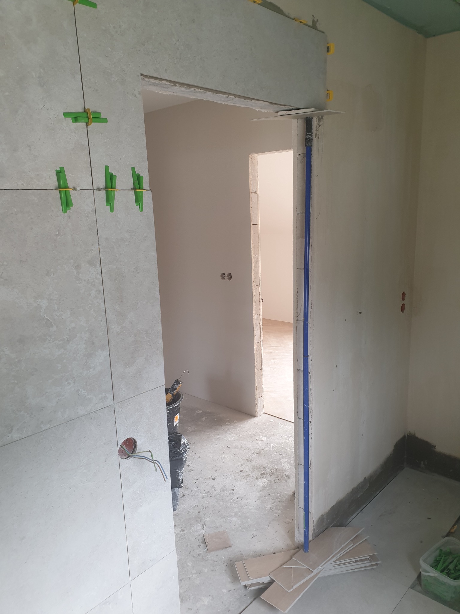 Montaż jasnych, kwadratowych płytek na ścianie z użyciem zielonych krzyżyków dystansowych. Widoczny otwór na gniazdko elektryczne i fragment rurki w ścianie.