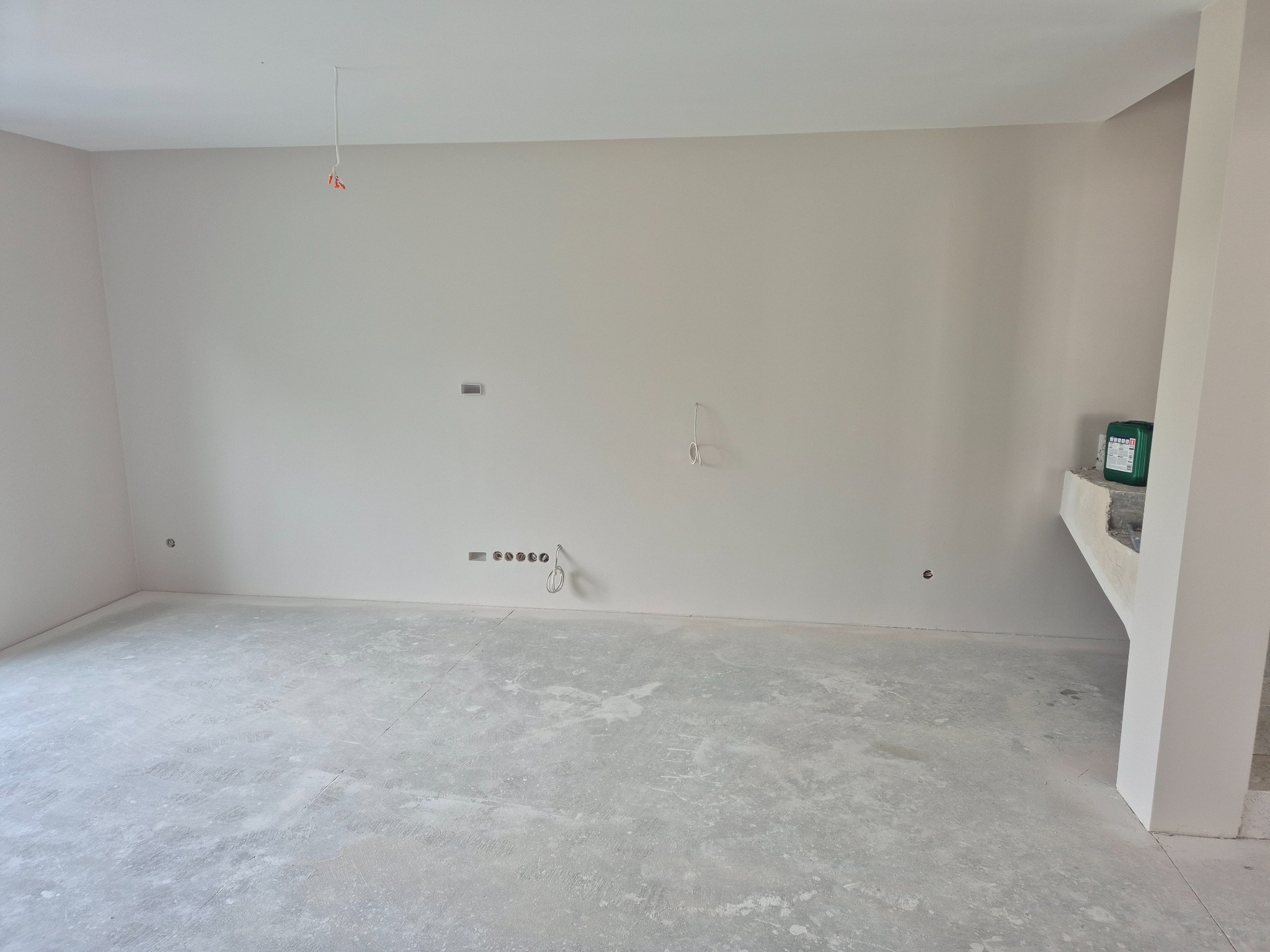 Jasne, puste wnętrze po szpachlowaniu ścian i wylewce podłogi, z widocznymi puszkami elektrycznymi i wystającymi przewodami. W rogu stoi zielony pojemnik.