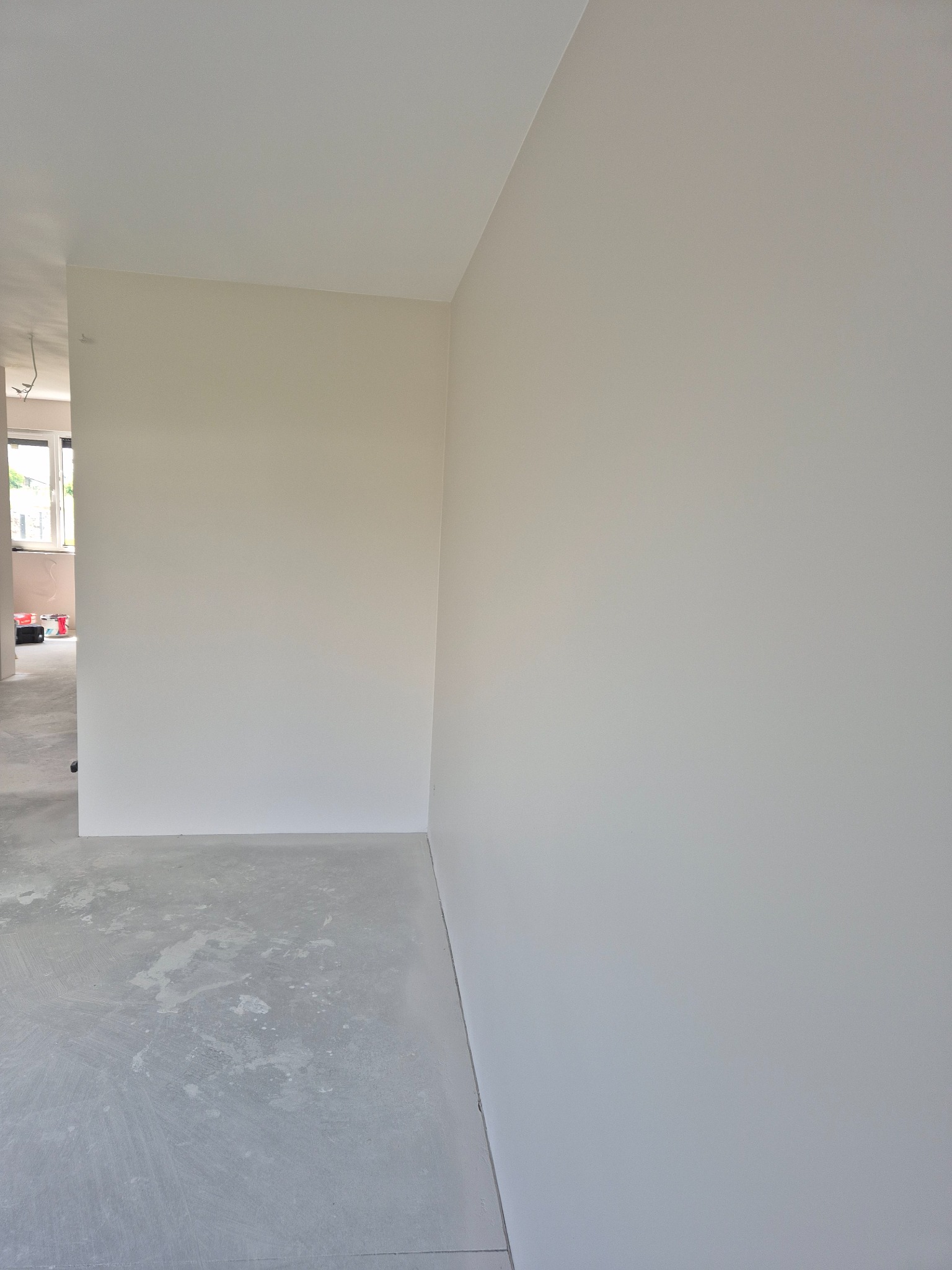 Wnętrze po szpachlowaniu i malowaniu na jasny odcień. Widoczne ślady na podłodze, okno w tle. Gładkie ściany w neutralnym oświetleniu.