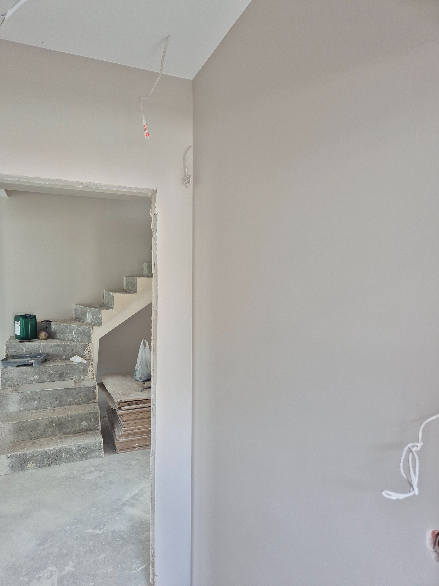 Świeżo pomalowana ściana w odcieniach szarości, z widocznymi schodami w tle. Widoczne kable elektryczne i materiały budowlane sugerują trwający remont.
