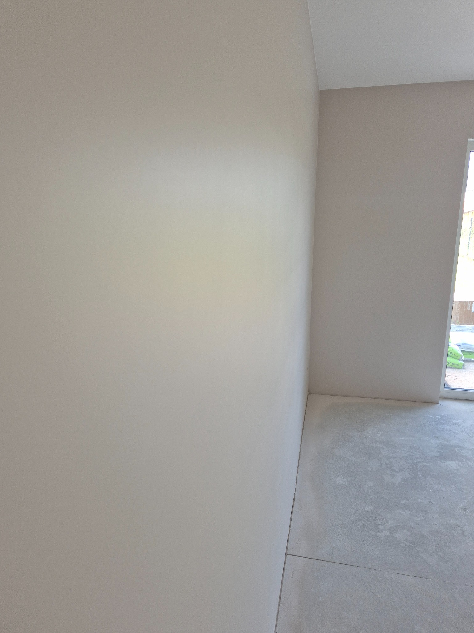 Świeżo pomalowane, jasnobeżowe ściany w pustym pokoju z oknem. Widoczna gładka powierzchnia ścian i surowa, betonowa podłoga. Minimalistyczny kadr, neutralne oświetlenie.