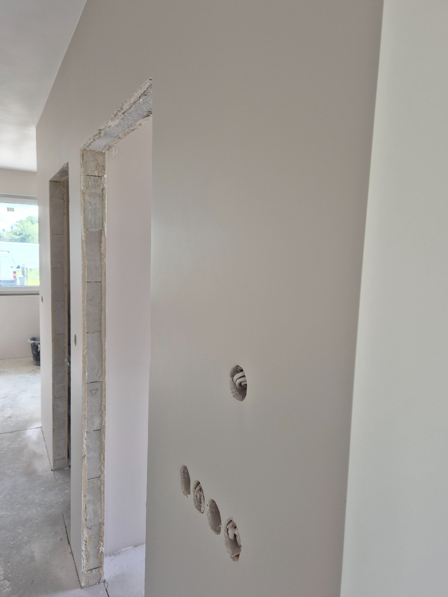 Wnętrze w trakcie wykańczania: gładkie, jasne ściany z otworami na gniazdka elektryczne. Widoczne surowe ościeżnice drzwiowe i fragment okna w tle.