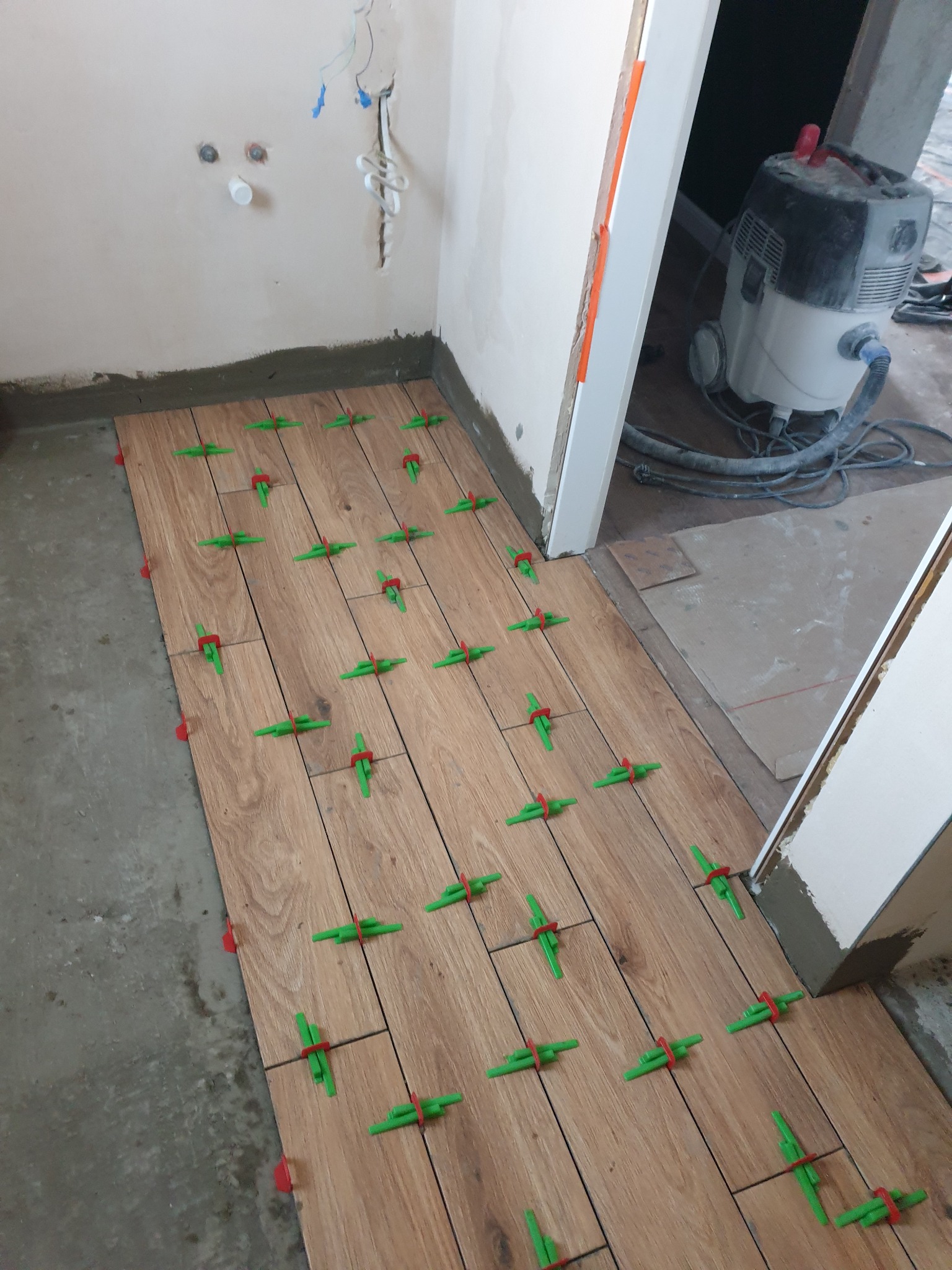 Układanie drewnopodobnych płytek podłogowych z użyciem zielonych krzyżyków dystansowych w trakcie remontu łazienki. W tle odkurzacz przemysłowy i ściany w trakcie prac.