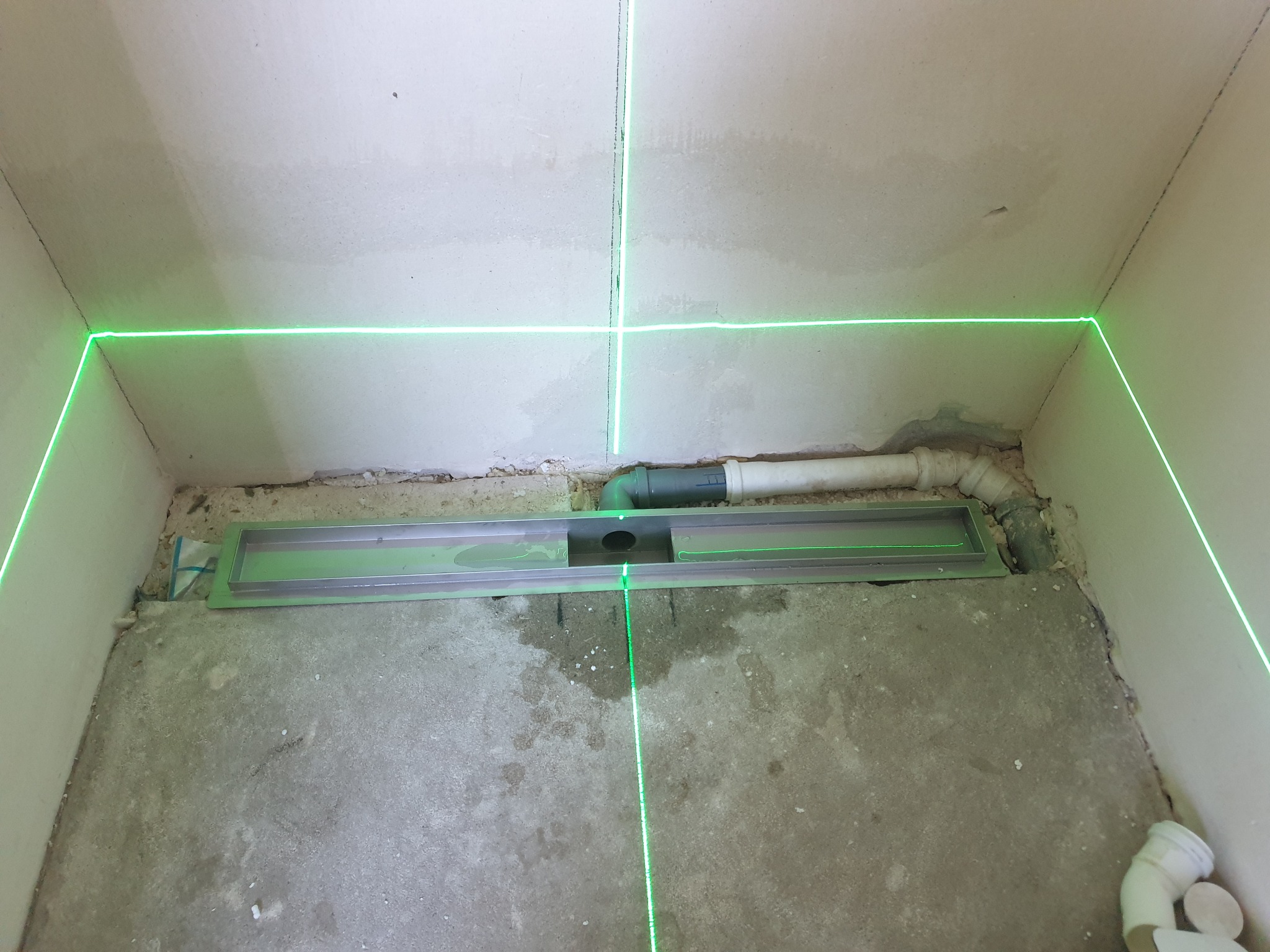 Montaż odpływu liniowego w łazience, wyznaczanie poziomu za pomocą lasera krzyżowego. Widoczne rury kanalizacyjne i surowy beton na podłodze.