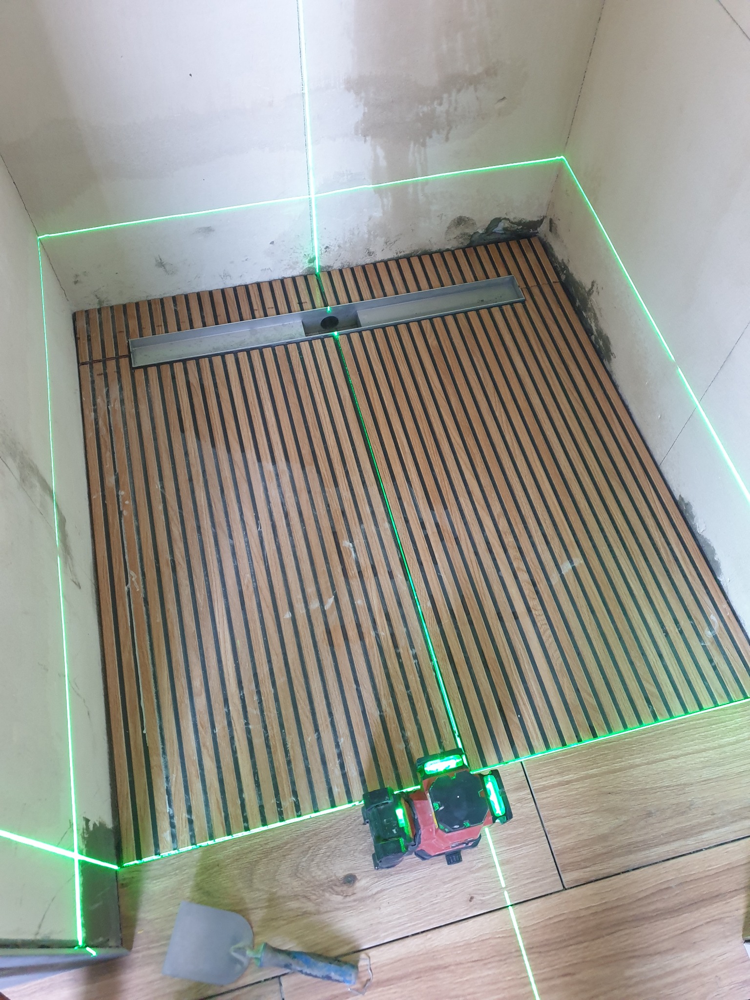 Ułożone deski w kabinie prysznicowej, wyznaczony poziomnicą laserową. Widoczna szpachelka, odpływ liniowy. Prace glazurnicze w trakcie realizacji.