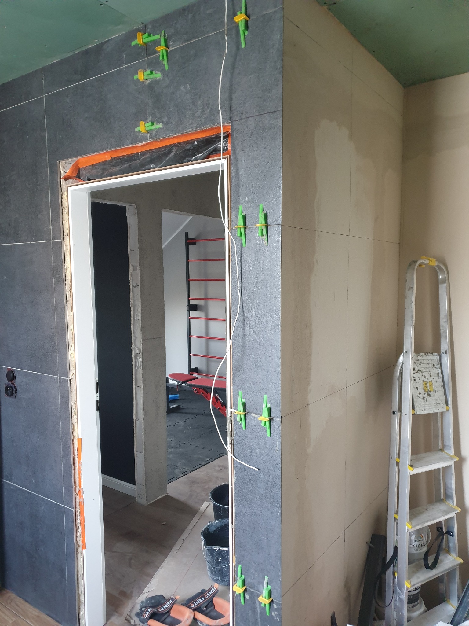 Montaż ciemnych płytek w łazience z użyciem klipsów poziomujących. W tle widoczna drabina i fragment pomieszczenia z drabinką gimnastyczną.