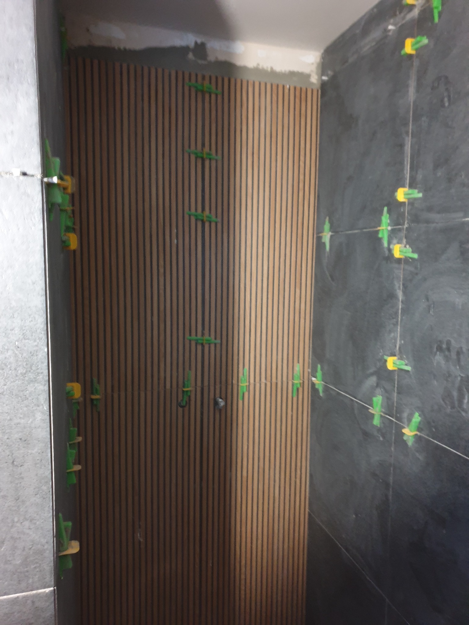 Montaż paneli dekoracyjnych i płytek w łazience z użyciem klipsów poziomujących. Widoczne pionowe panele drewnopodobne i ciemne płytki.