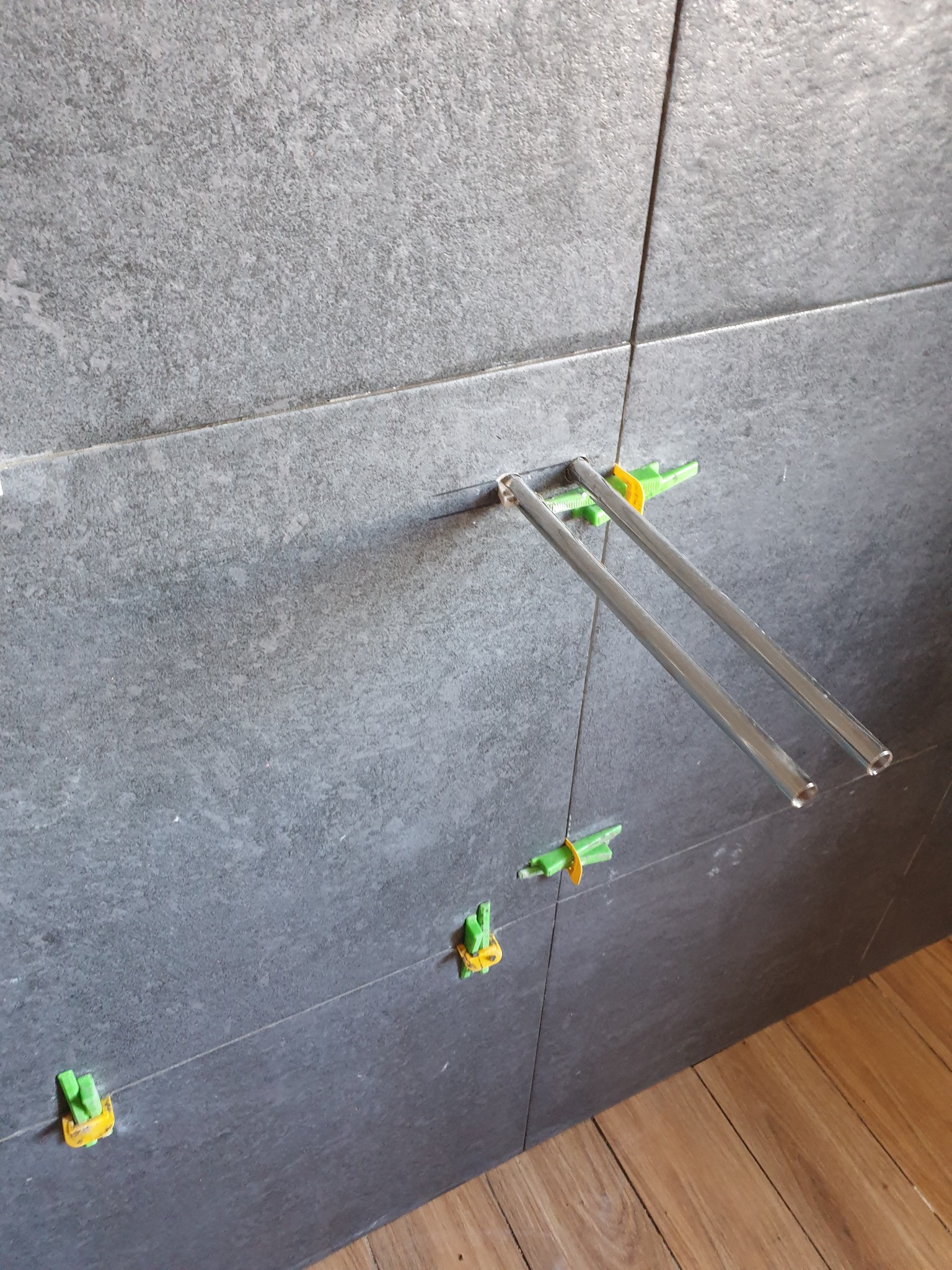 Montaż szarych płytek z użyciem systemu poziomowania, widoczne metalowe pręty i zielono-żółte klipsy dystansowe. Podłoga z paneli imitujących drewno.