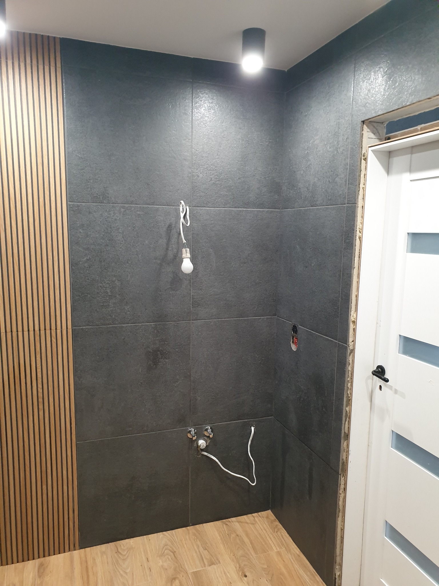 Szare płytki w łazience z drewnianą ścianką, widoczna instalacja wodna i żarówka na kablu. Drzwi białe z przeszkleniami. Oświetlenie sufitowe.