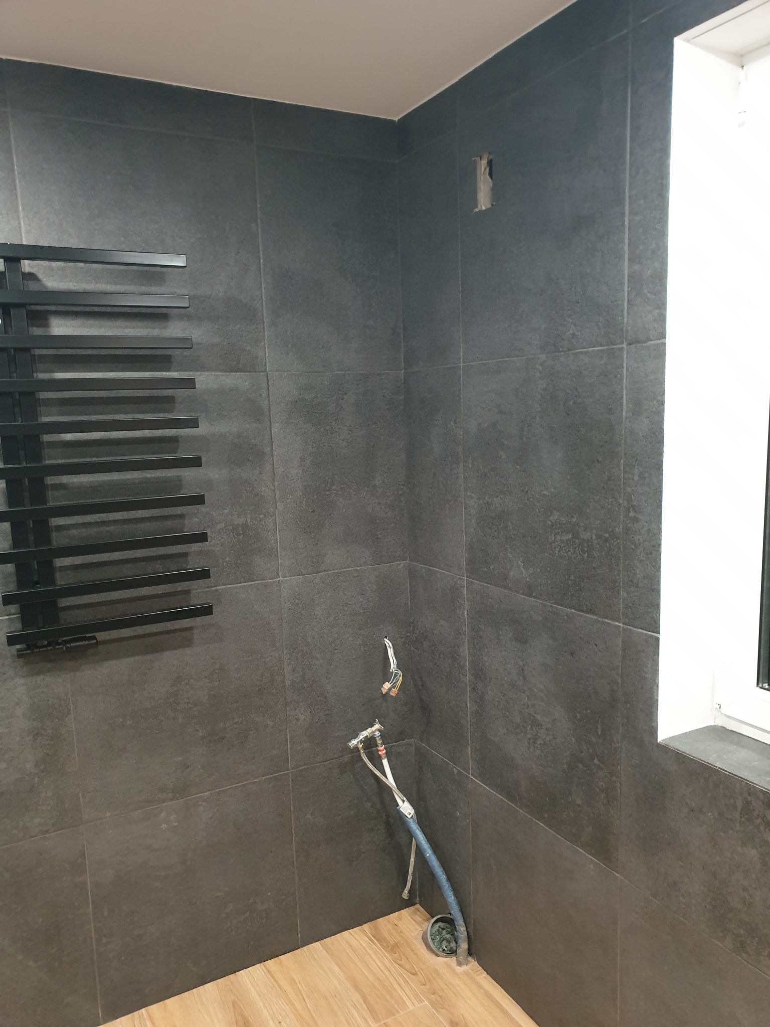 Ciemne, matowe płytki ścienne w trakcie montażu w łazience. Grzejnik drabinkowy, instalacja wodna i okno widoczne. Podłoga wyłożona panelami.