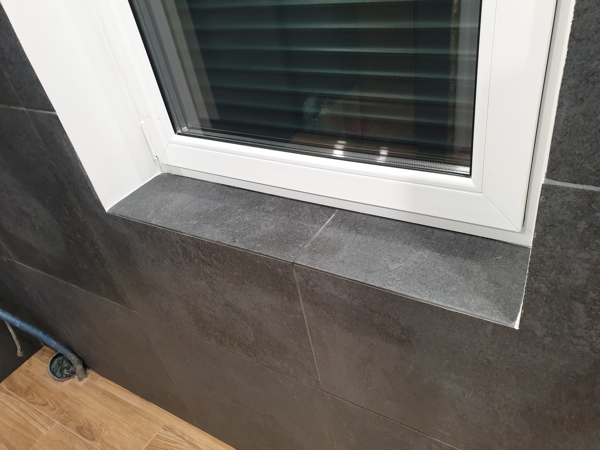 Szare płytki ścienne i biała rama okienna w łazience w Sapowicach. Minimalistyczne wykończenie z widocznym odpływem podłogowym i drewnopodobną podłogą.