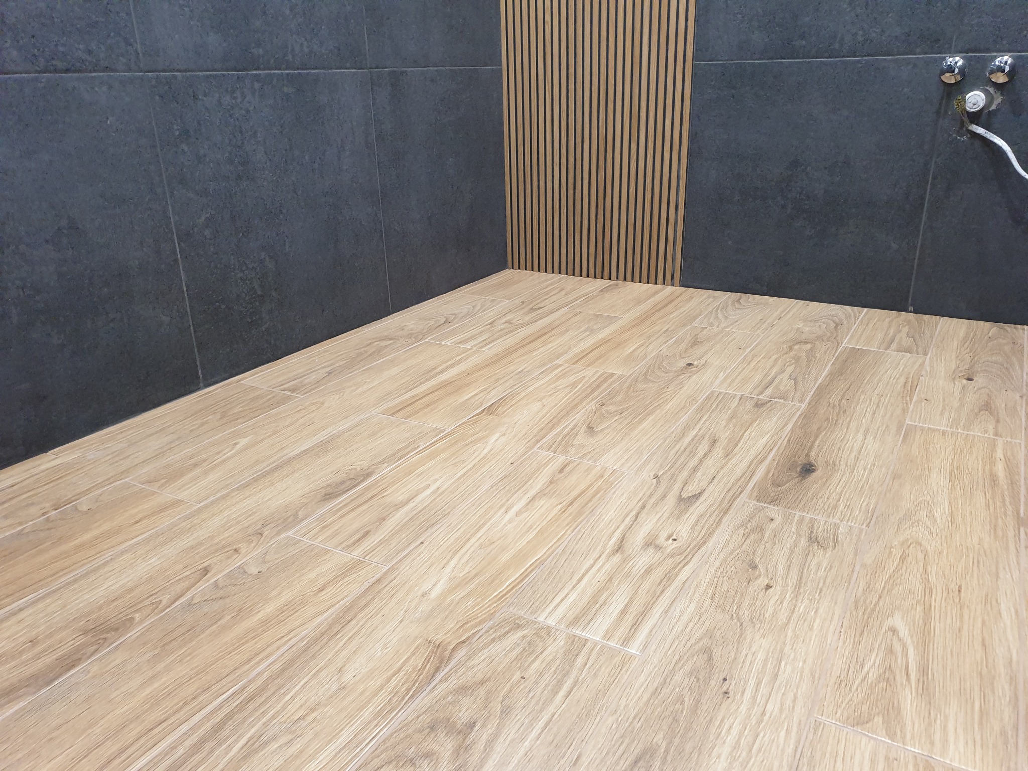 Wykończony narożnik łazienki z drewnopodobną podłogą, ciemnymi płytkami ściennymi i dekoracyjnym panelem z drewnianych listewek. Widoczne przyłącza armatury.