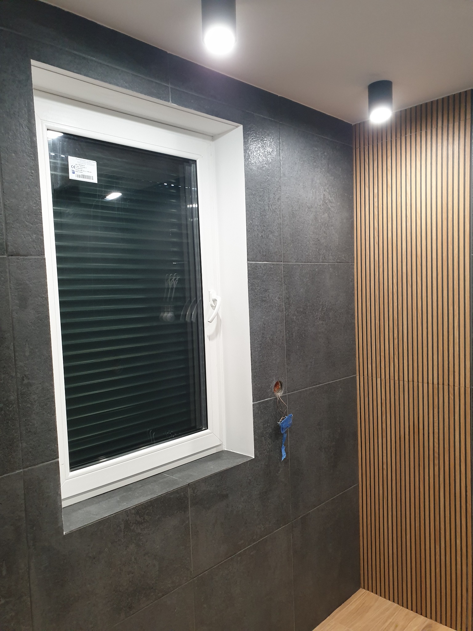 Nowoczesna łazienka z szarą, teksturowaną glazurą, drewnianymi lamelami na ścianie i białym oknem z żaluzjami plisowanymi. Widoczne oświetlenie sufitowe.