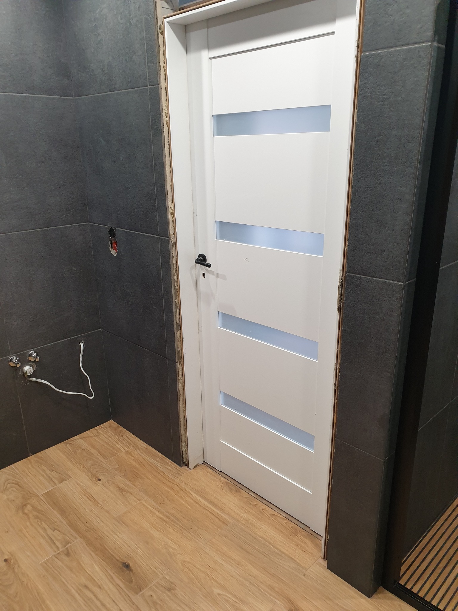 Wykończona łazienka z drewnopodobną podłogą i ciemnymi płytkami ściennymi. Białe drzwi z przeszkleniami, widoczne instalacje wodne i elektryczne.