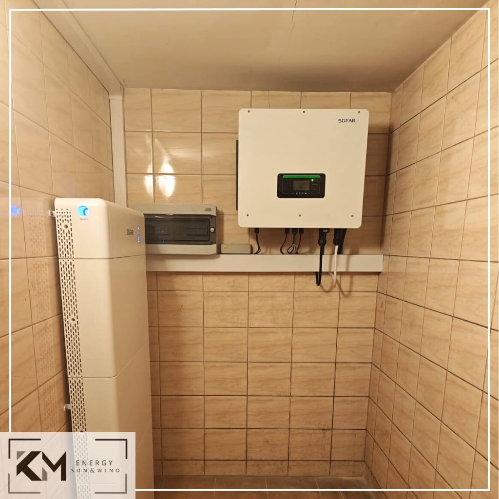 Biały akumulator energii, falownik i skrzynka elektryczna zamontowane na ścianie z beżowych płytek w pomieszczeniu.