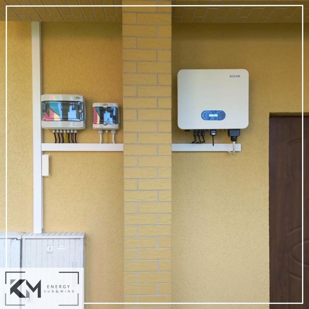 Instalacja fotowoltaiczna na elewacji budynku: falownik SOFAR i dwie skrzynki z bezpiecznikami, z widocznymi przewodami i skrzynką dolną. Dodatkowo widoczne logo firmy KM Energy Sun&Wind w lewym...