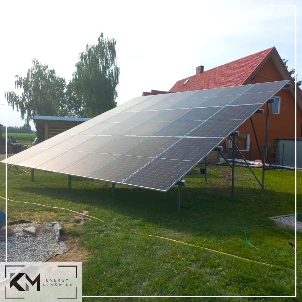 Instalacja paneli słonecznych na metalowej konstrukcji na trawniku przed pomarańczowym domem z czerwoną dachówką, widoczne drzewa w tle, logo firmy w lewym dolnym rogu.