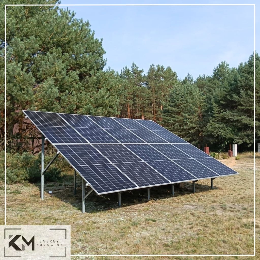 Instalacja paneli słonecznych na metalowej konstrukcji, widok na las iglasty w tle, logo firmy w rogu zdjęcia.