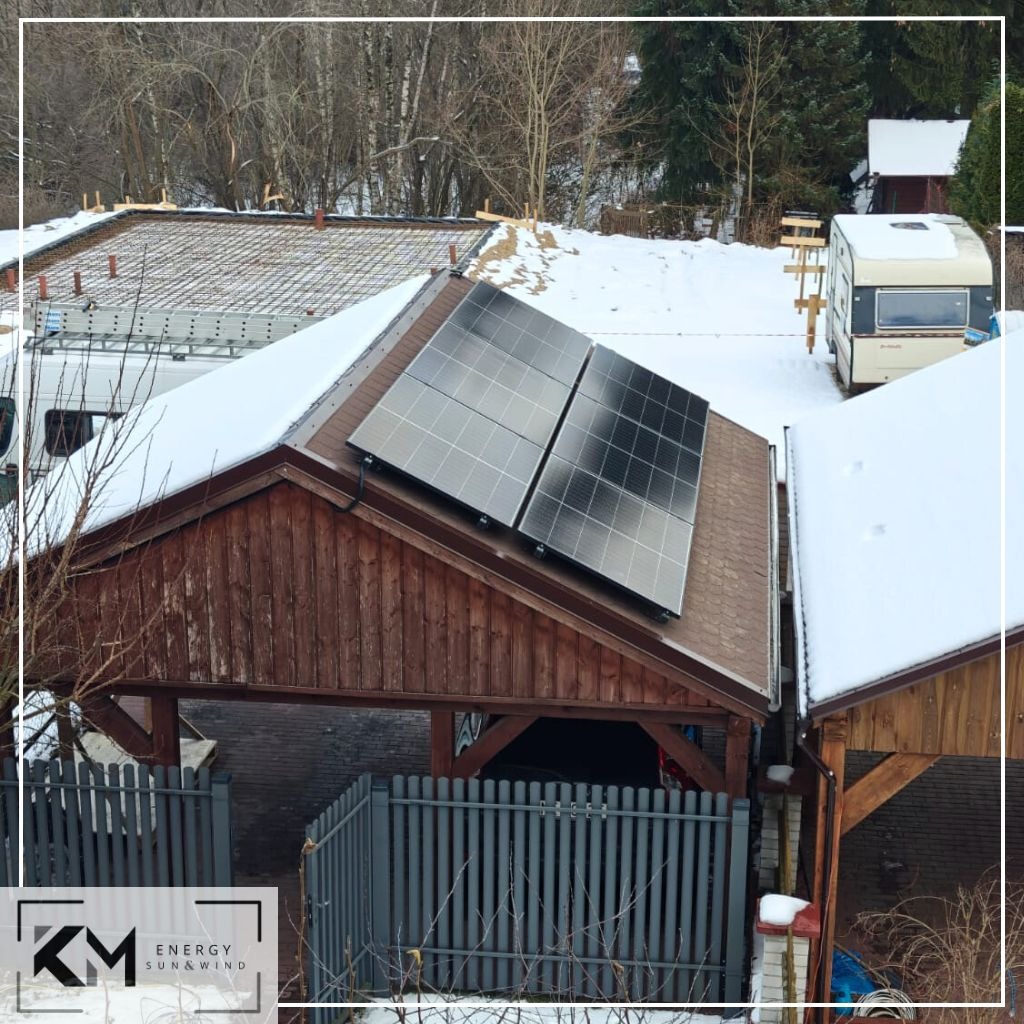 Panele słoneczne zamontowane na dachu drewnianej wiaty garażowej w zimowej scenerii, częściowo pokryte śniegiem, widoczne z góry, z logo firmy w rogu.