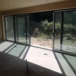 Wnętrze w stanie surowym z gładkimi ścianami i sufitem po nałożeniu tynku. Widoczne duże okno przesuwne z wyjściem na zewnątrz, gdzie leżą deski i stoi drabina.