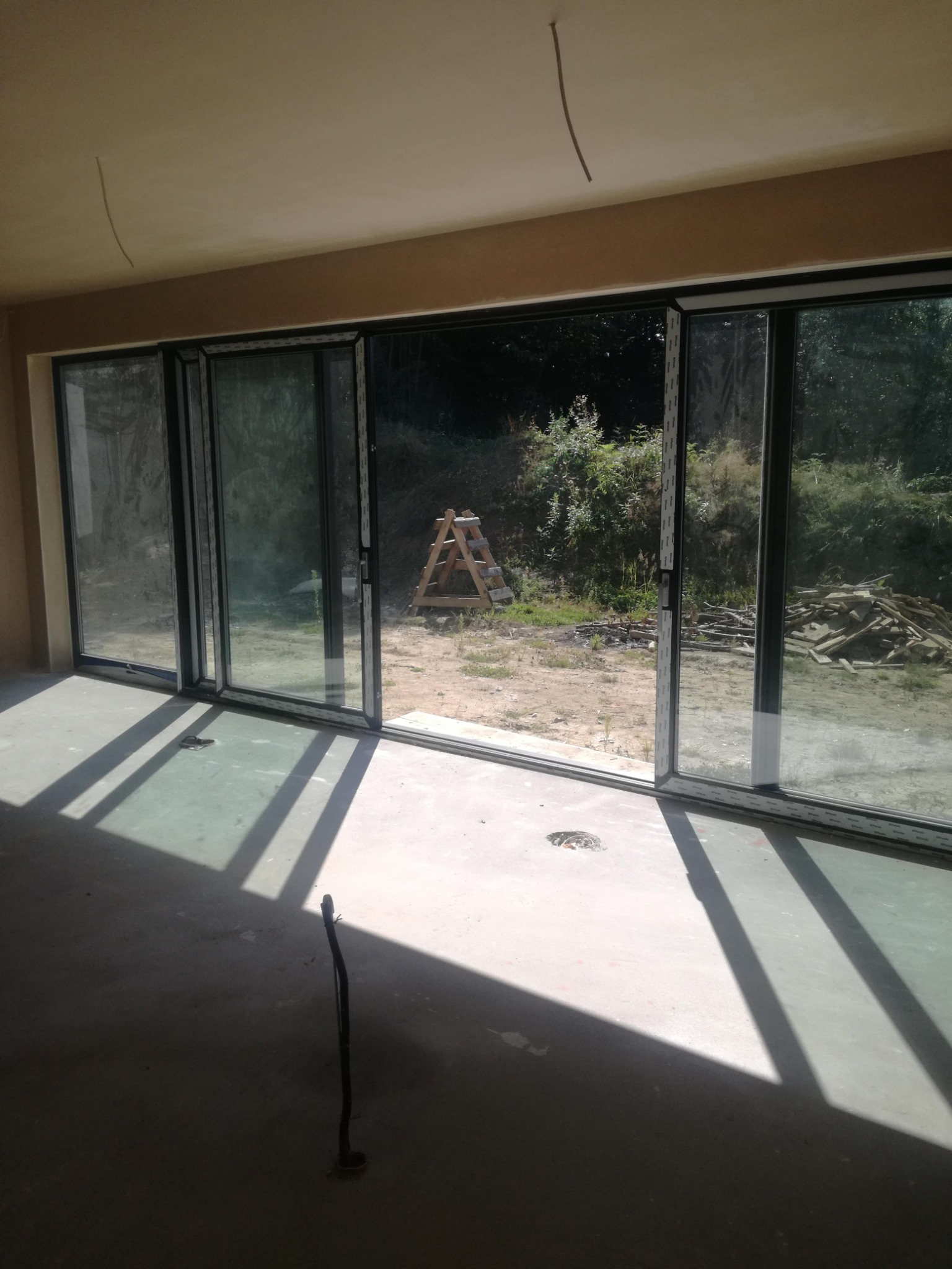 Wnętrze w stanie surowym z gładkimi ścianami i sufitem po nałożeniu tynku. Widoczne duże okno przesuwne z wyjściem na zewnątrz, gdzie leżą deski i stoi drabina.