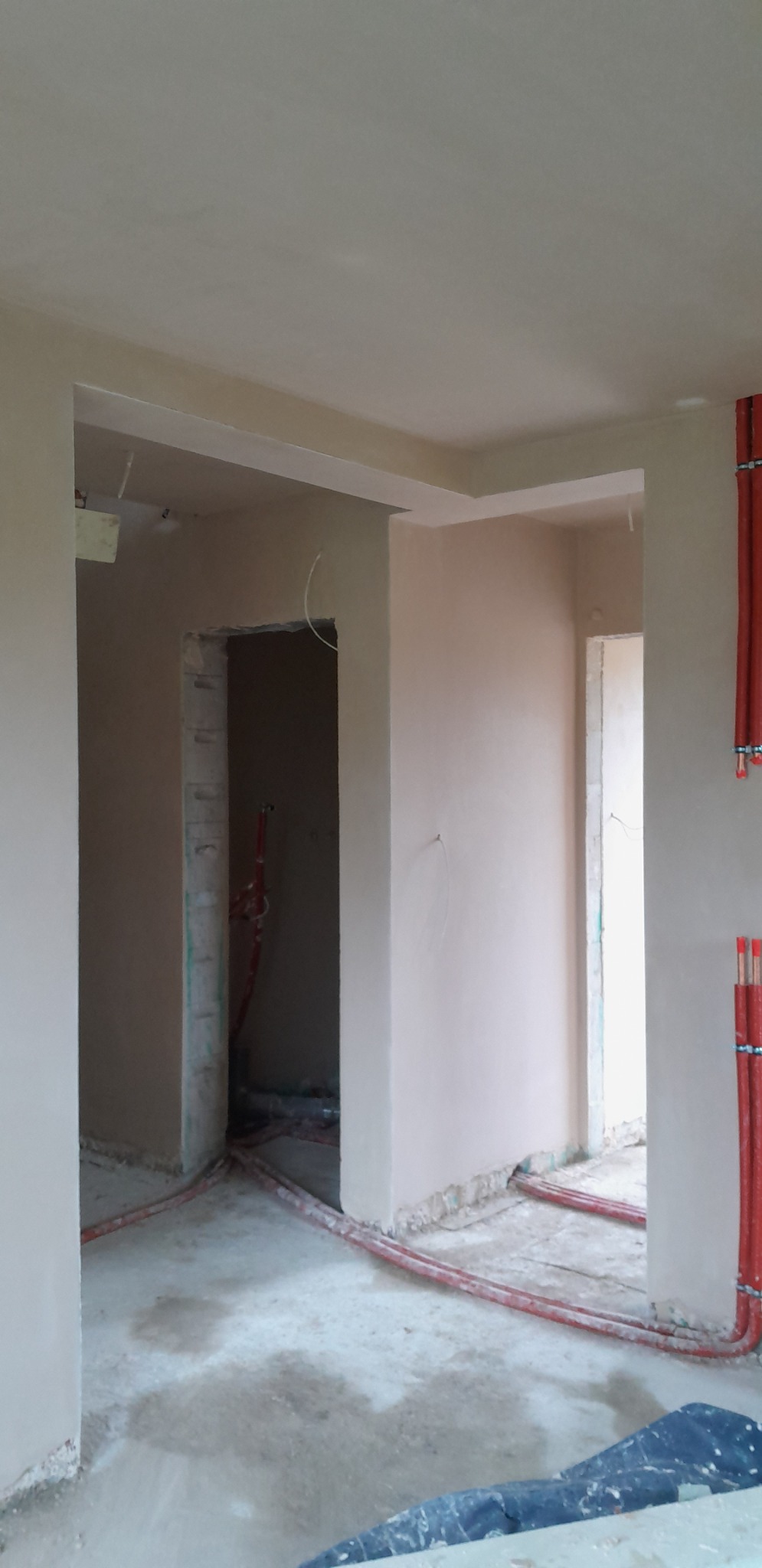 Wnętrze po nałożeniu tynków: gładkie, beżowe ściany, widoczne instalacje grzewcze w czerwonych rurach, sufit z elementami dekoracyjnymi. Stan surowy, przygotowany do dalszych prac.
