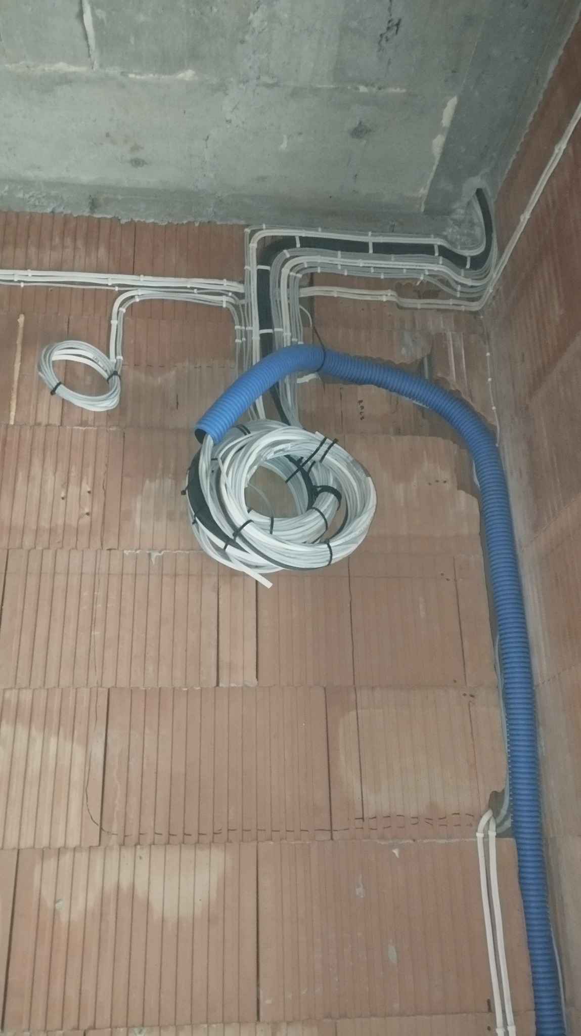 Surowe wnętrze w trakcie prac: kable elektryczne prowadzone po ceglanej ścianie i betonowym suficie, wiązka kabli zwinięta w okrąg, niebieska rura karbowana.