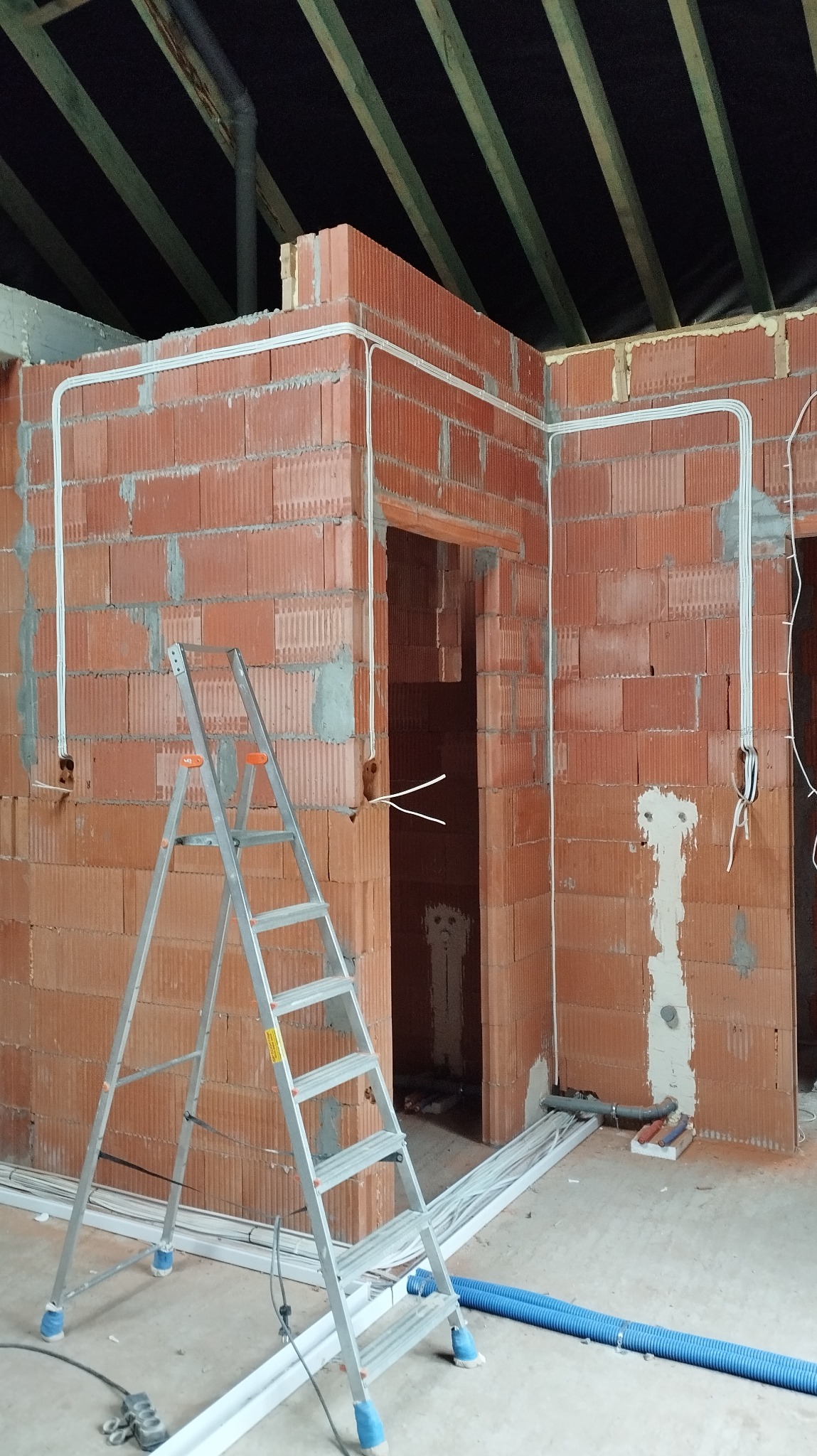 Instalacja elektryczna w budynku w trakcie budowy, kable prowadzone po ścianach z czerwonej cegły, drabina aluminiowa, peszle na podłodze.