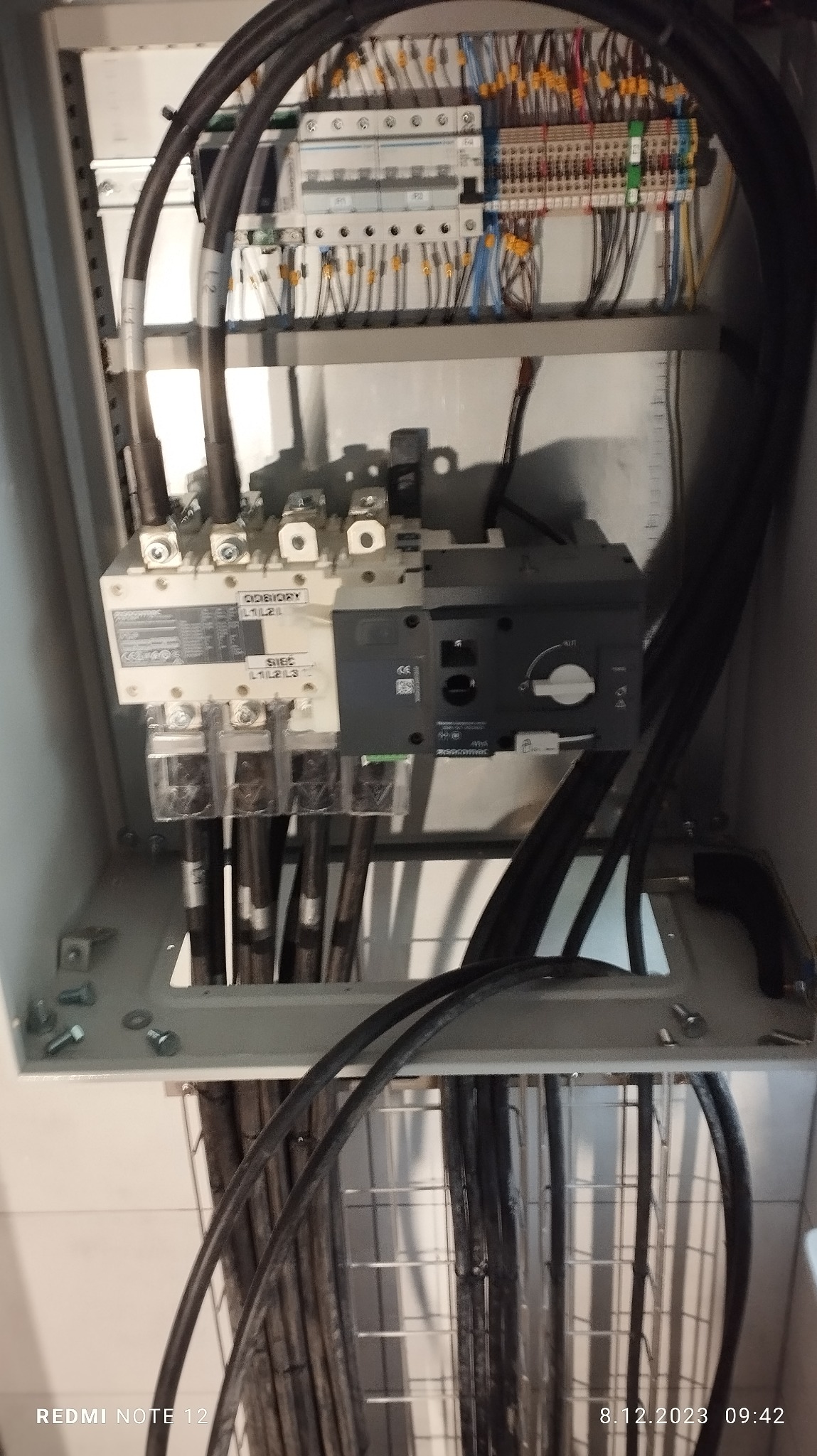 Otwarta szafa elektryczna z widocznymi zabezpieczeniami, przewodami i listwami zaciskowymi; widoczne śruby montażowe.