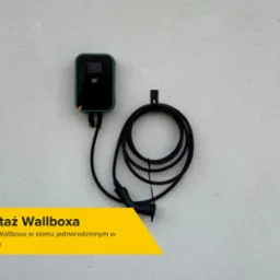 Czarny wallbox marki GC zamontowany na jasnej, zewnętrznej ścianie budynku, kabel zwinięty w pętlę, widoczny uchwyt mocujący.