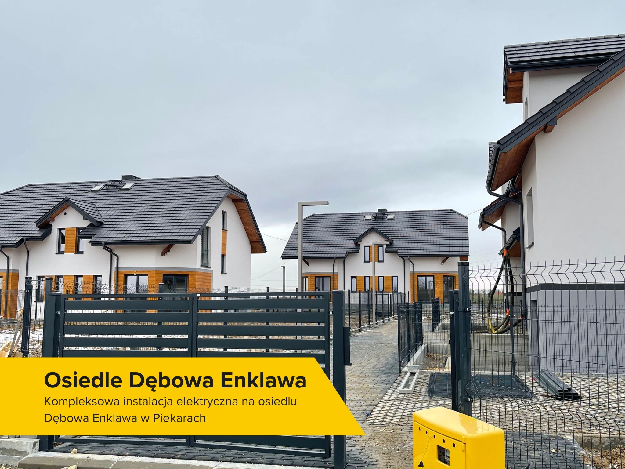 Nowoczesne osiedle domów jednorodzinnych z szarą dachówką i grafitowymi ogrodzeniami panelowymi, widoczna skrzynka elektryczna w kolorze żółtym oraz kable wystające z elewacji jednego z domów.