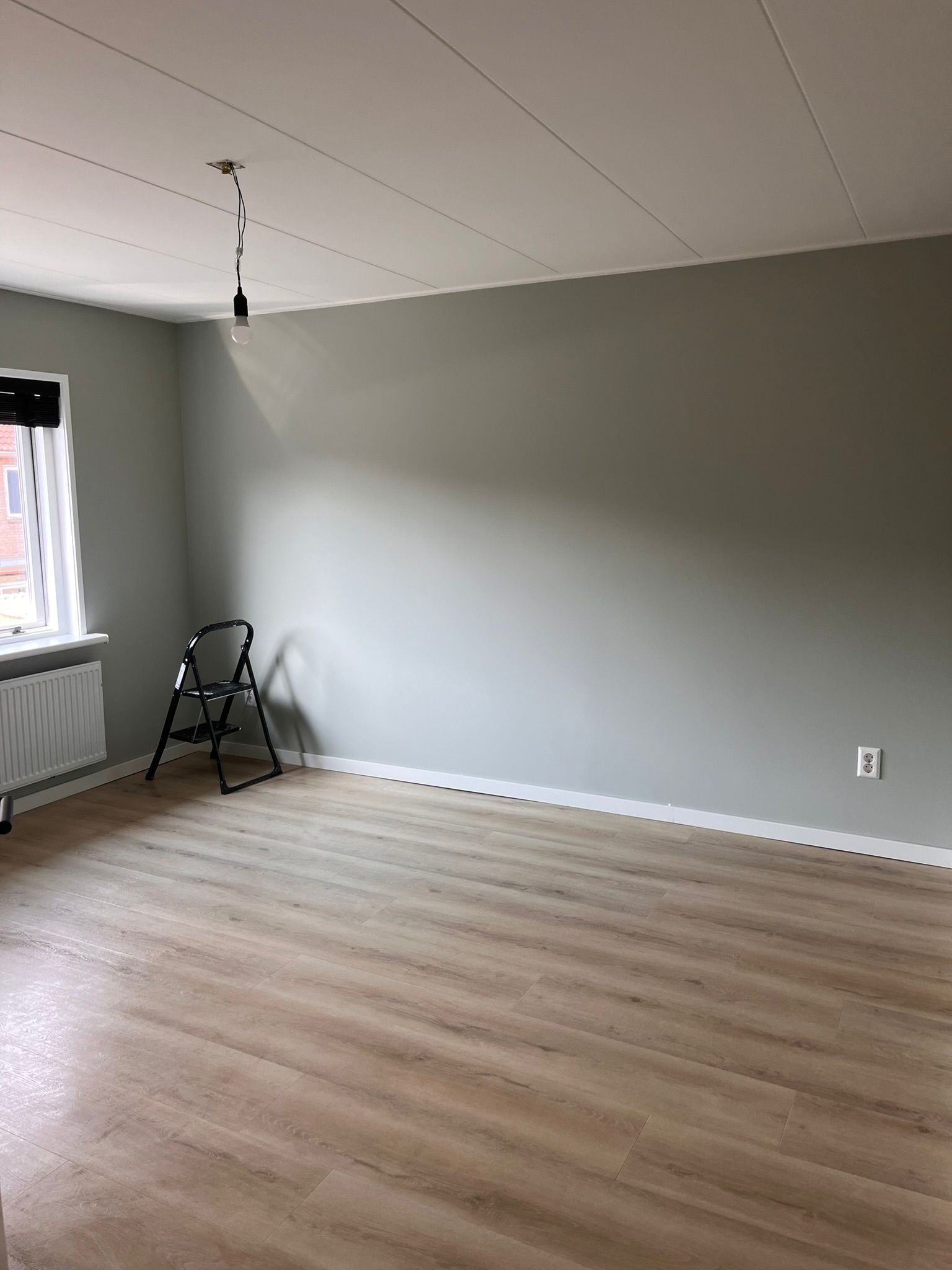 Pomalowane na szaro puste pomieszczenie z nową podłogą z paneli i drabinką malarską, widok na okno z grzejnikiem i wiszącą żarówkę.