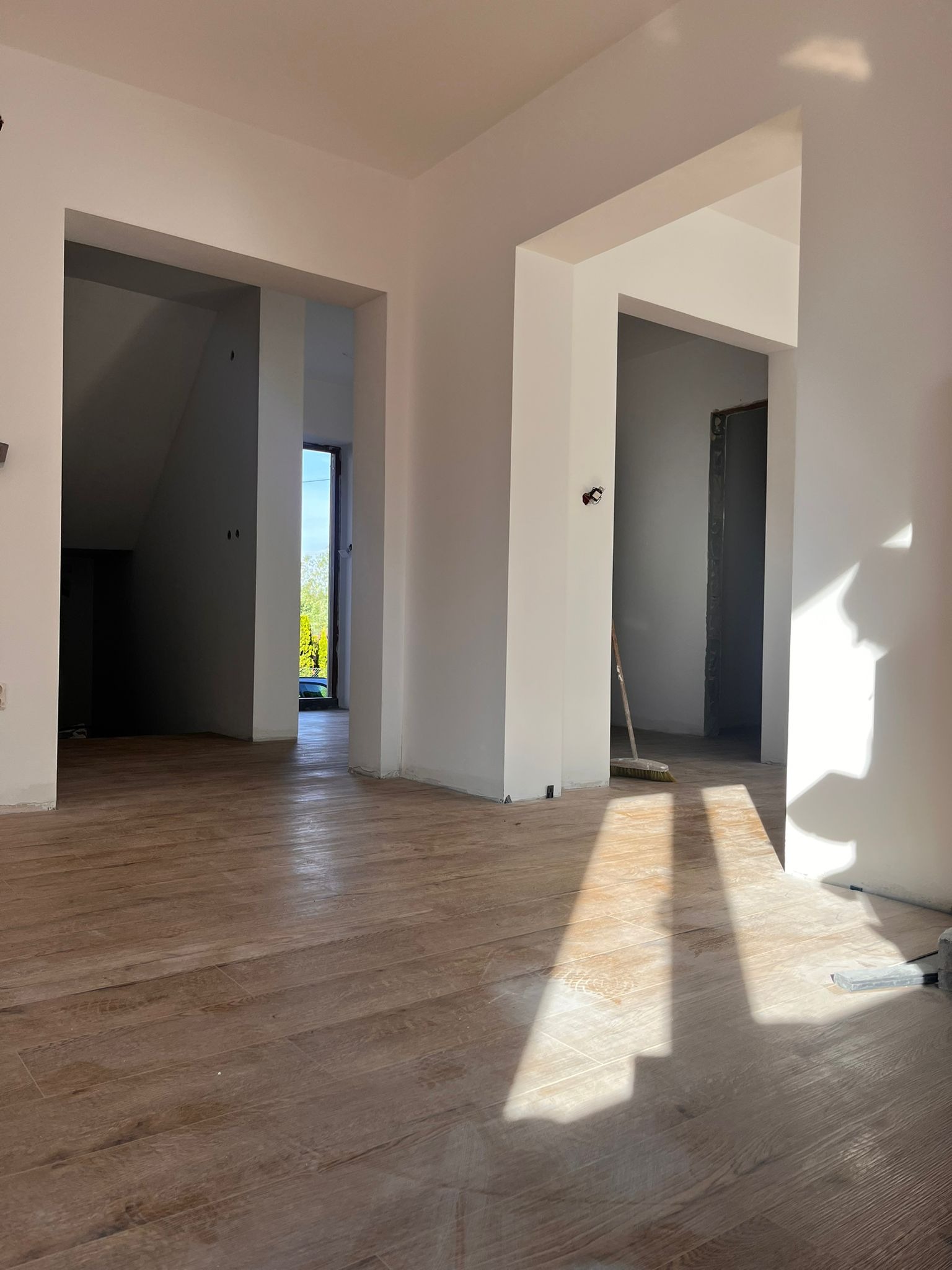 Wnętrze w trakcie prac wykończeniowych: świeżo pomalowane ściany na biało, drewnopodobne panele podłogowe, widoczne ościeżnice i cień słoneczny na podłodze, w tle miotła.