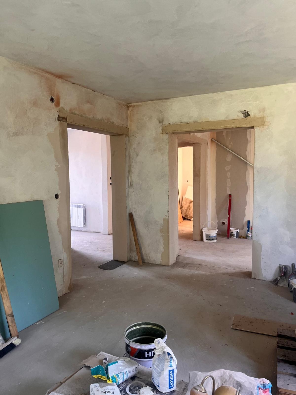 Wnętrze w trakcie prac przygotowawczych: surowe ściany po nałożeniu warstwy szpachli, widoczne otwarte przejścia do kolejnych pomieszczeń, na podłodze wiadro z farbą i worki z zaprawą, pionowo...