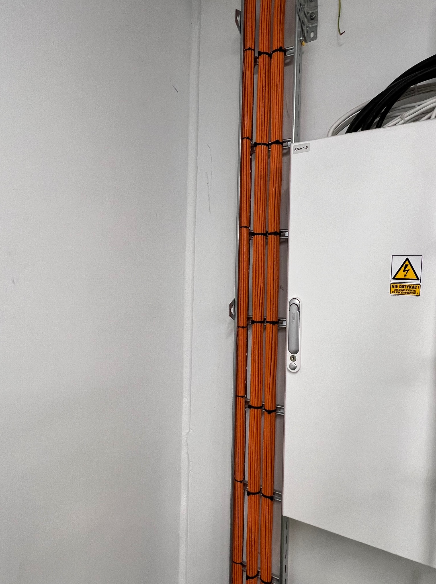 Pomarańczowe kable sieciowe ułożone równolegle w metalowej korycie, przymocowane czarnymi opaskami zaciskowymi, biegnące wzdłuż białej szafy elektrycznej z ostrzeżeniem o wysokim napięciu.