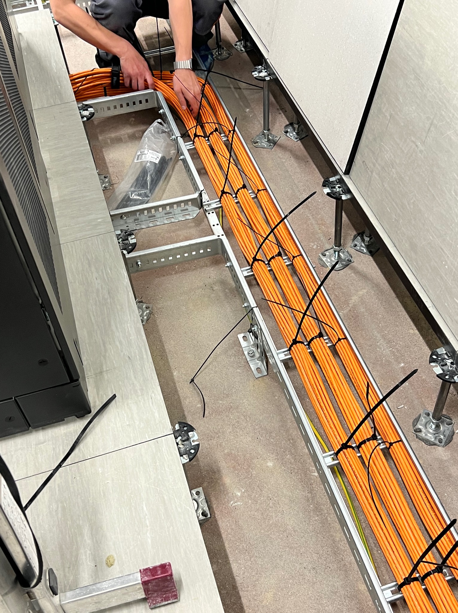 Instalacja pomarańczowych kabli sieciowych w korycie pod podłogą techniczną, zabezpieczonych czarnymi opaskami zaciskowymi; widoczny fragment szafy rack i ręce instalatora w szarym dresie.