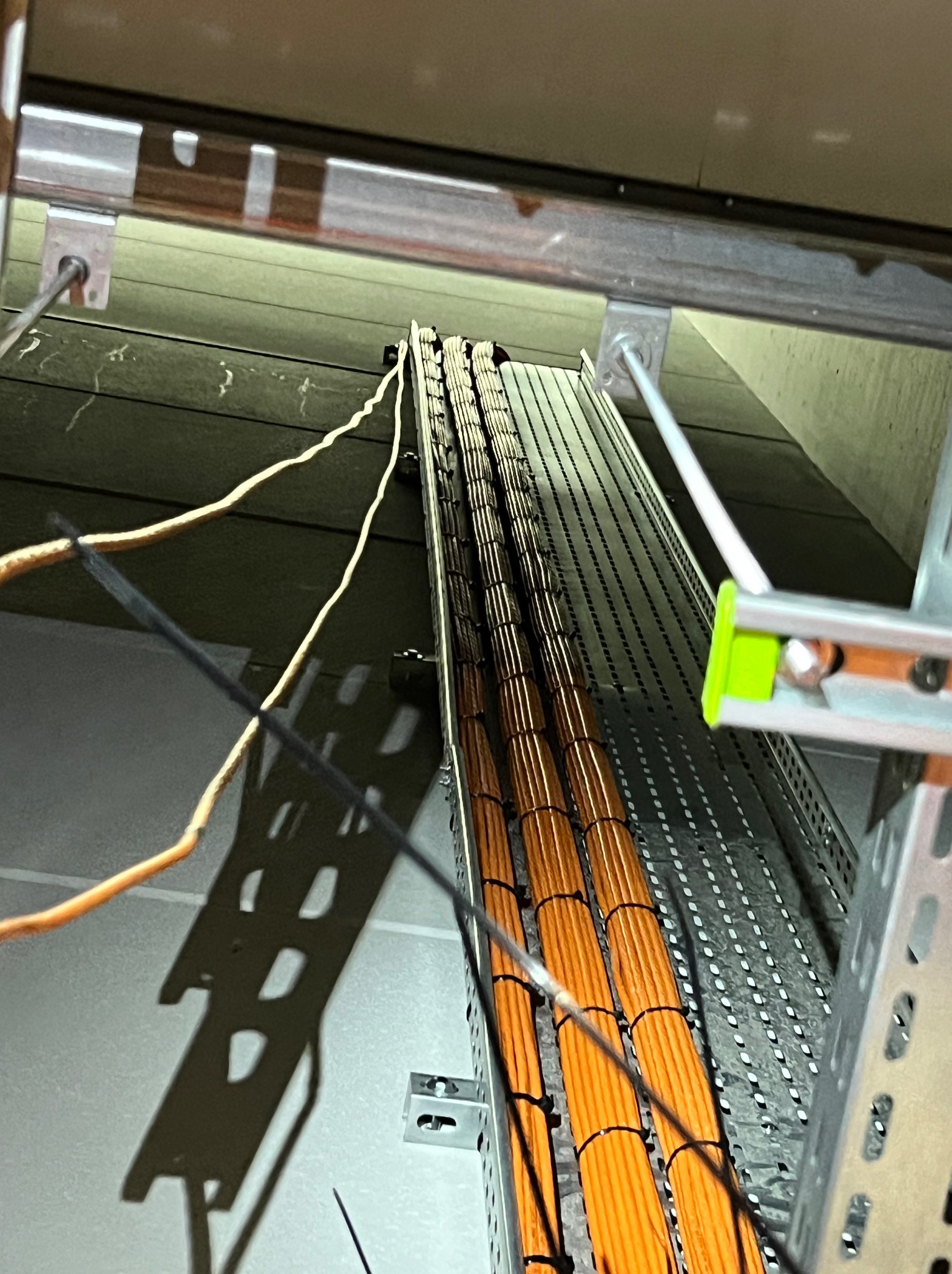 Pomarańczowe kable ułożone w metalowej korycie kablowej pod sufitem, widoczne elementy mocujące i pojedyncze, luźne przewody.