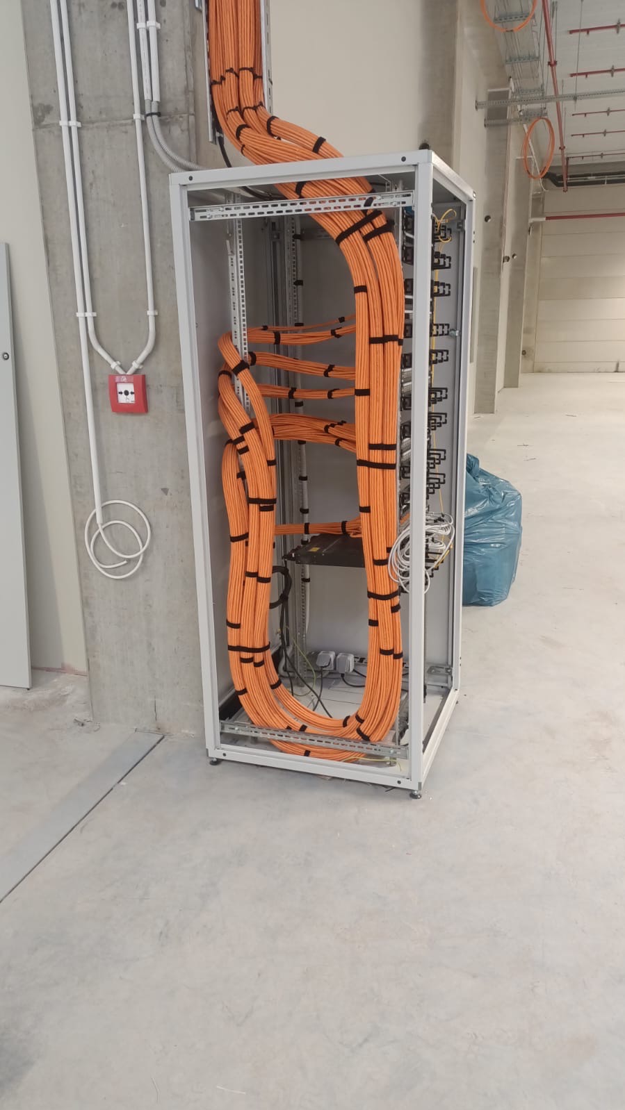 Pomarańczowe przewody starannie ułożone w szafie rack, zabezpieczone czarnymi opaskami, na tle surowego, betonowego wnętrza z widocznymi instalacjami pod sufitem.
