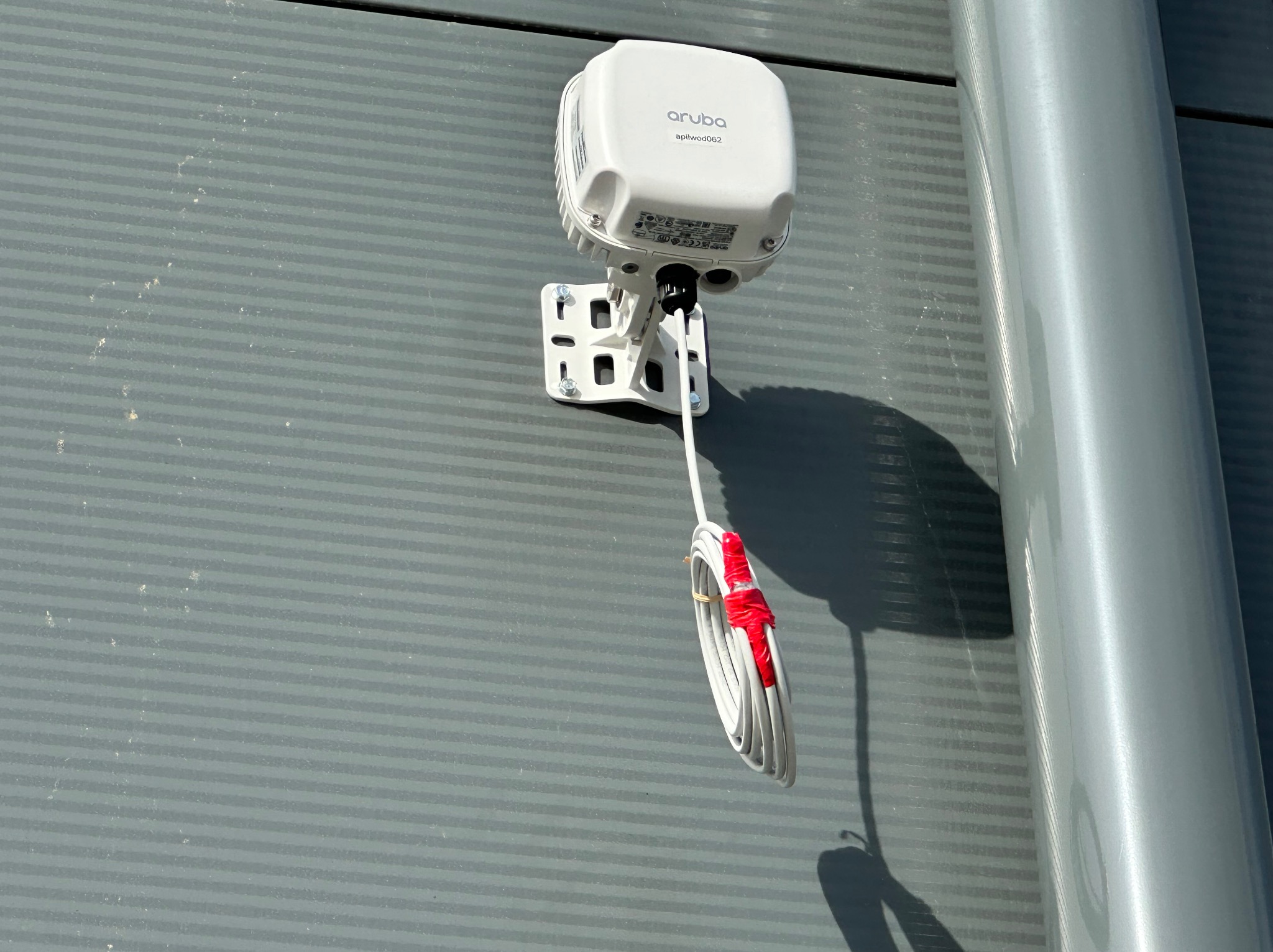 Biały punkt dostępowy Aruba zamontowany na szarej ścianie z widocznym zwiniętym kablem zabezpieczonym czerwoną taśmą.