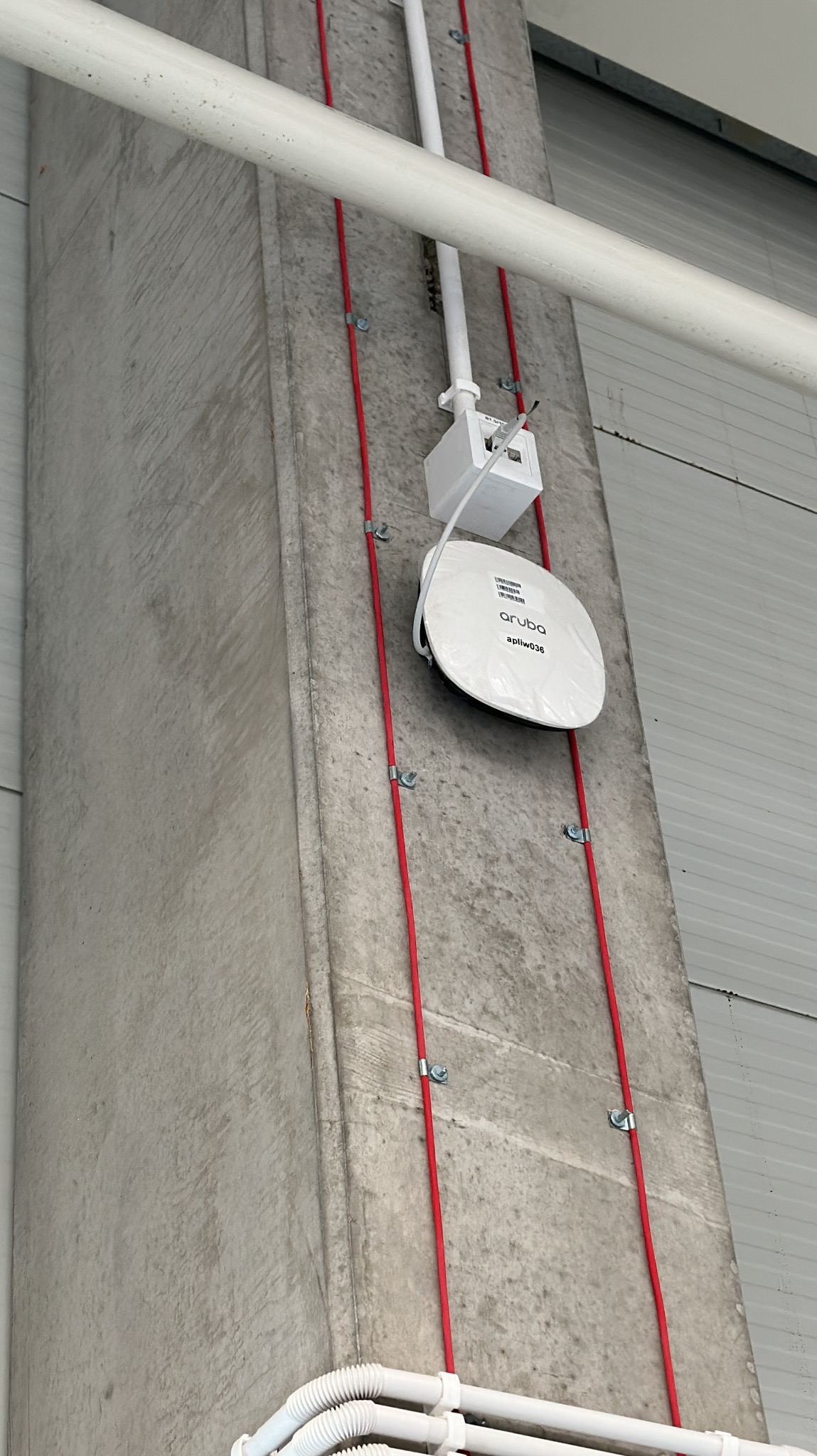 Biały punkt dostępowy Aruba apliw036 zamontowany na betonowym słupie, z widocznymi czerwonymi kablami przymocowanymi metalowymi uchwytami i białymi rurami instalacyjnymi.