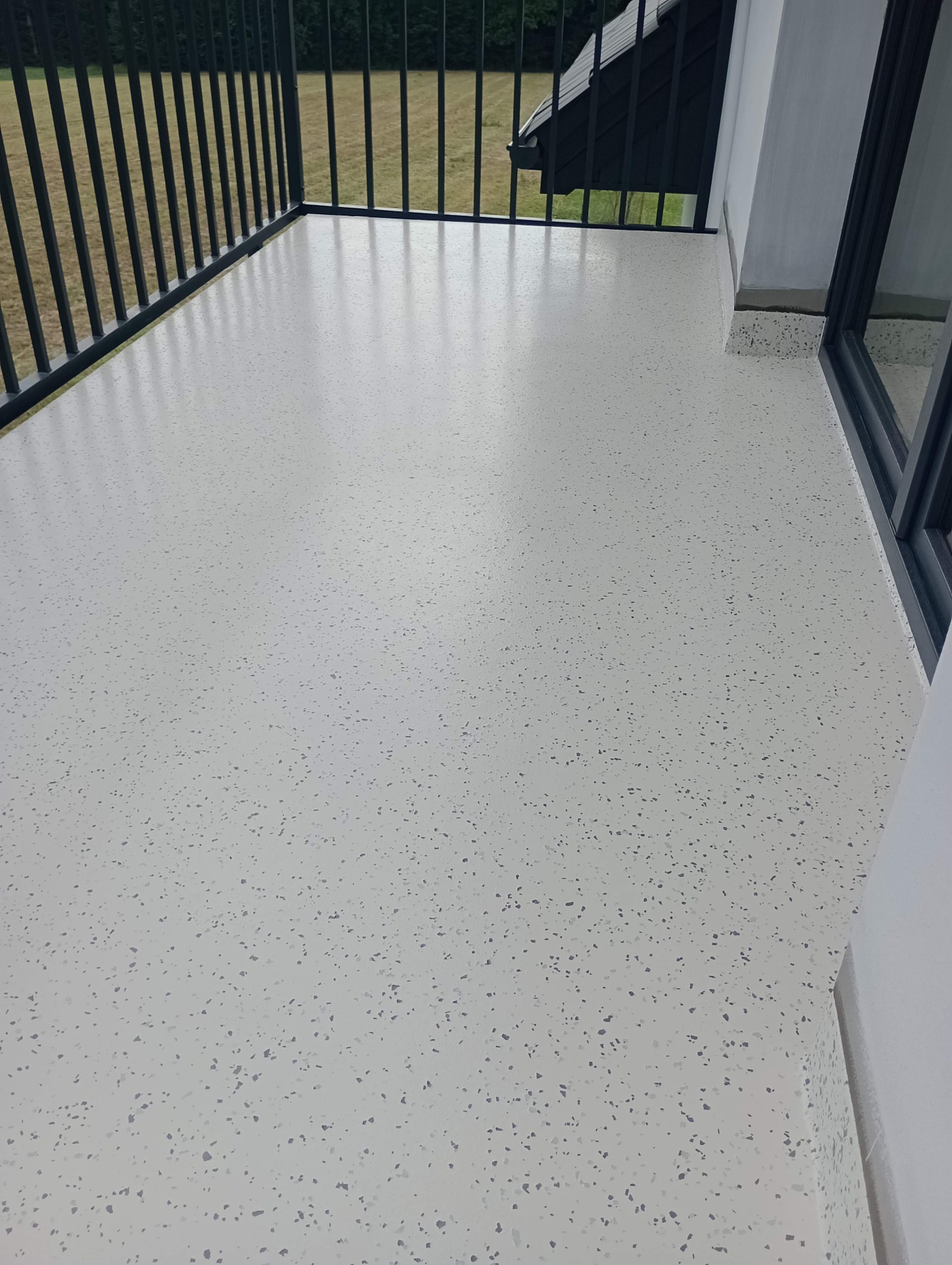 Jasnoszara posadzka żywiczna z drobnymi, ciemniejszymi plamkami na balkonie z czarną, metalową balustradą i widokiem na trawnik.