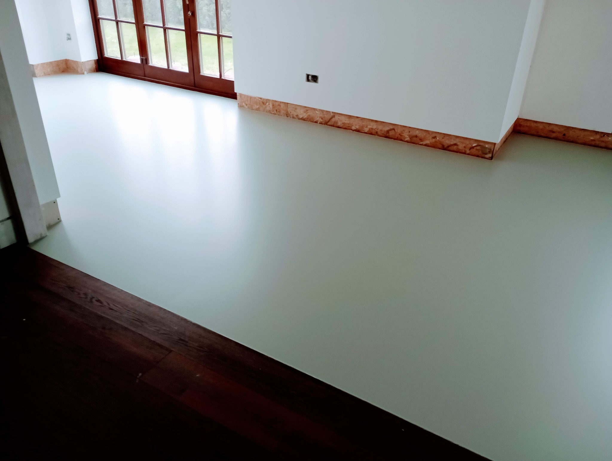 Jasnoszara, gładka posadzka w jasnym pokoju z oknami balkonowymi, widoczne przygotowane ściany do wykończenia.