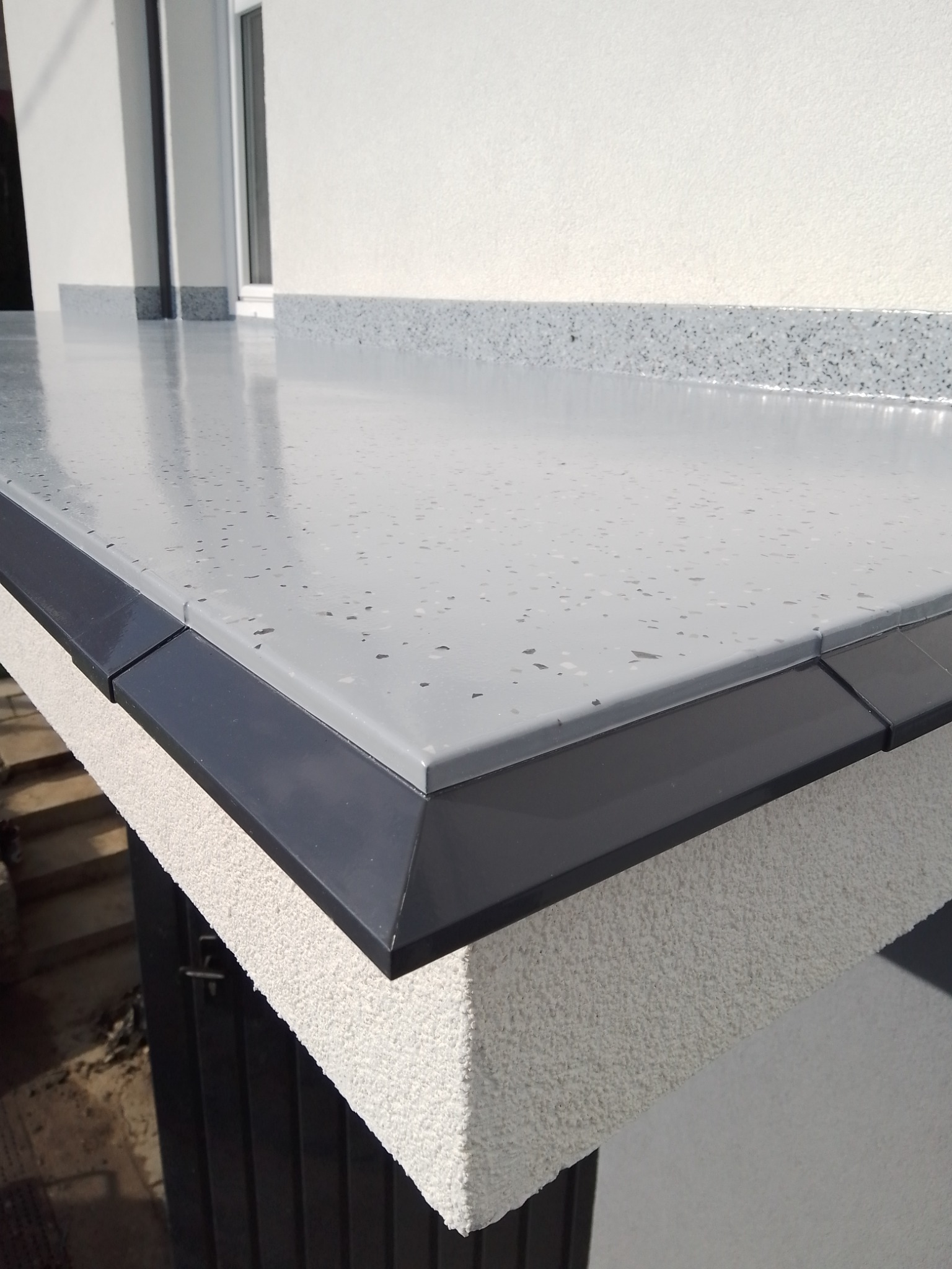 Szara, błyszcząca powierzchnia balkonu wykończona czarnym obrzeżem, z widoczną fakturą ściany i fragmentem okna w tle.