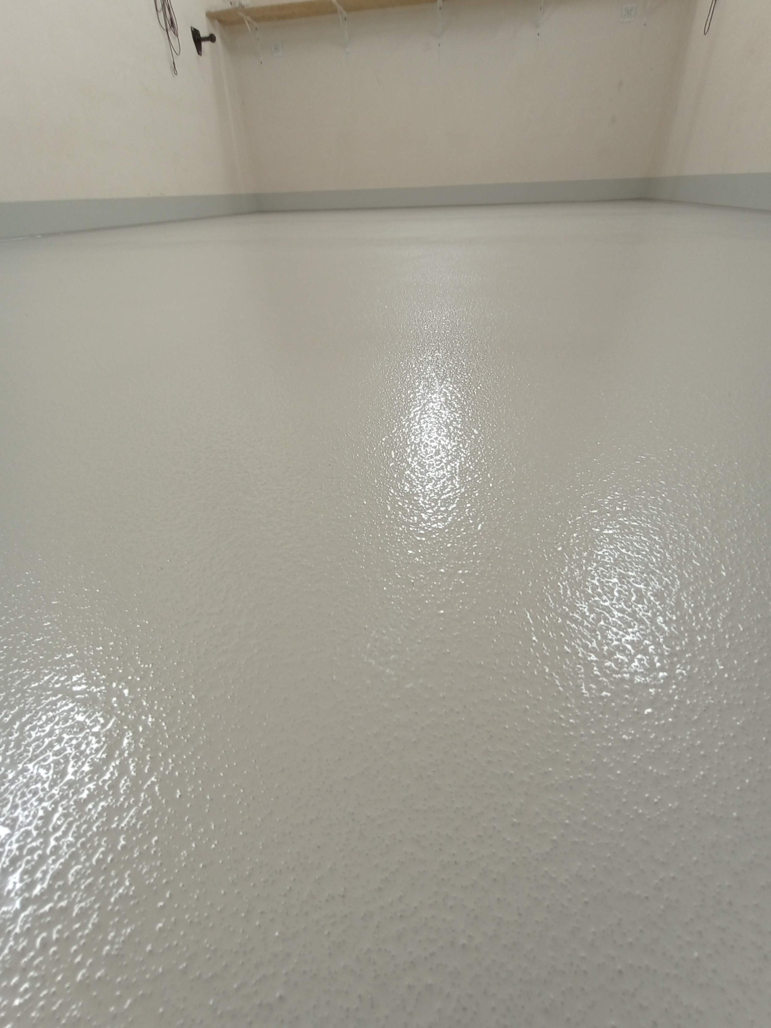 Jasnoszara, gładka posadzka żywiczna z delikatną fakturą, świeżo wylana, widok z niskiej perspektywy, przy listwie przypodłogowej, w tle biała ściana i półka.